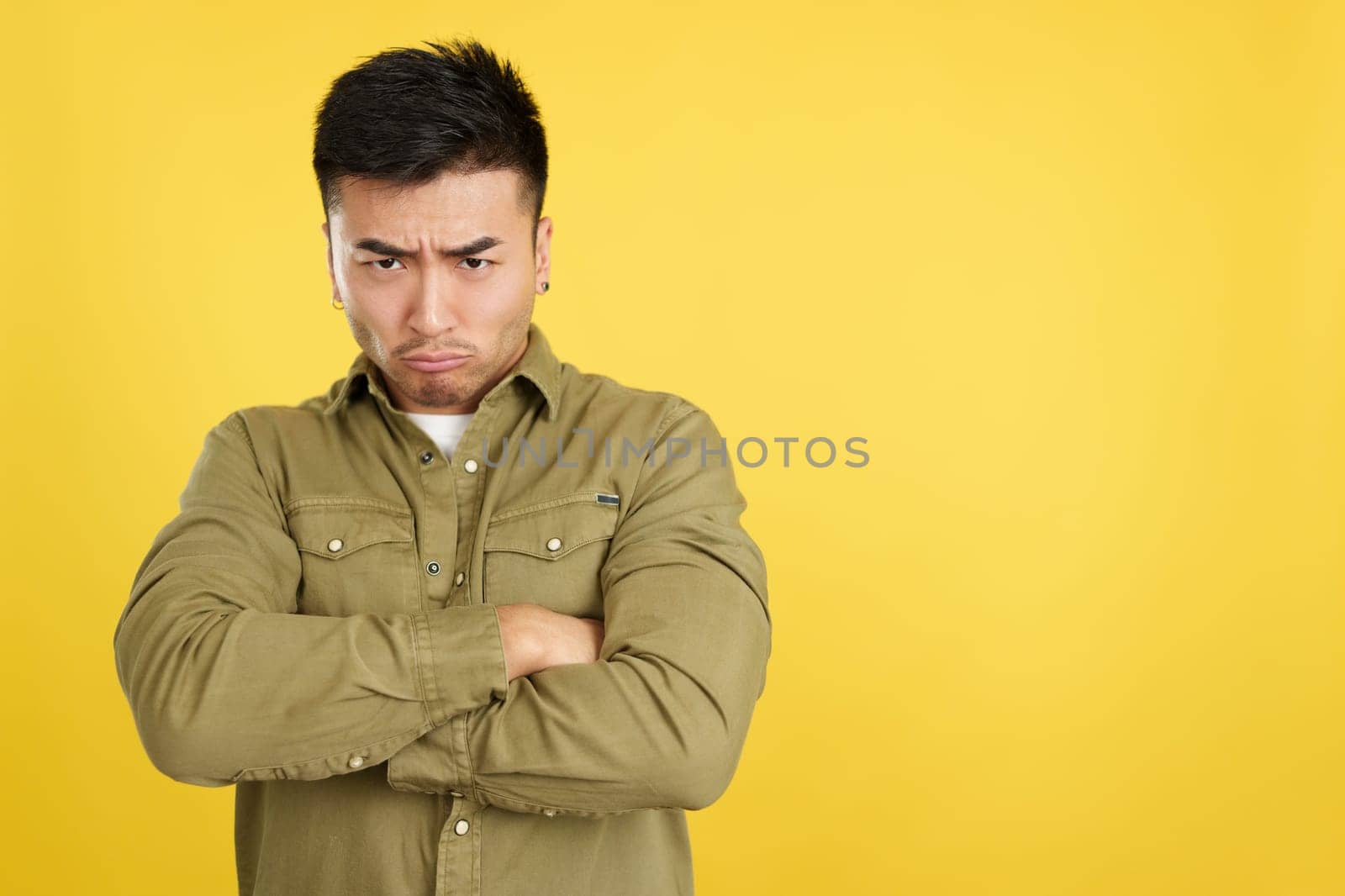 Chinese man crossing the arms with an angry expression while looking at camera in studio with yellow background