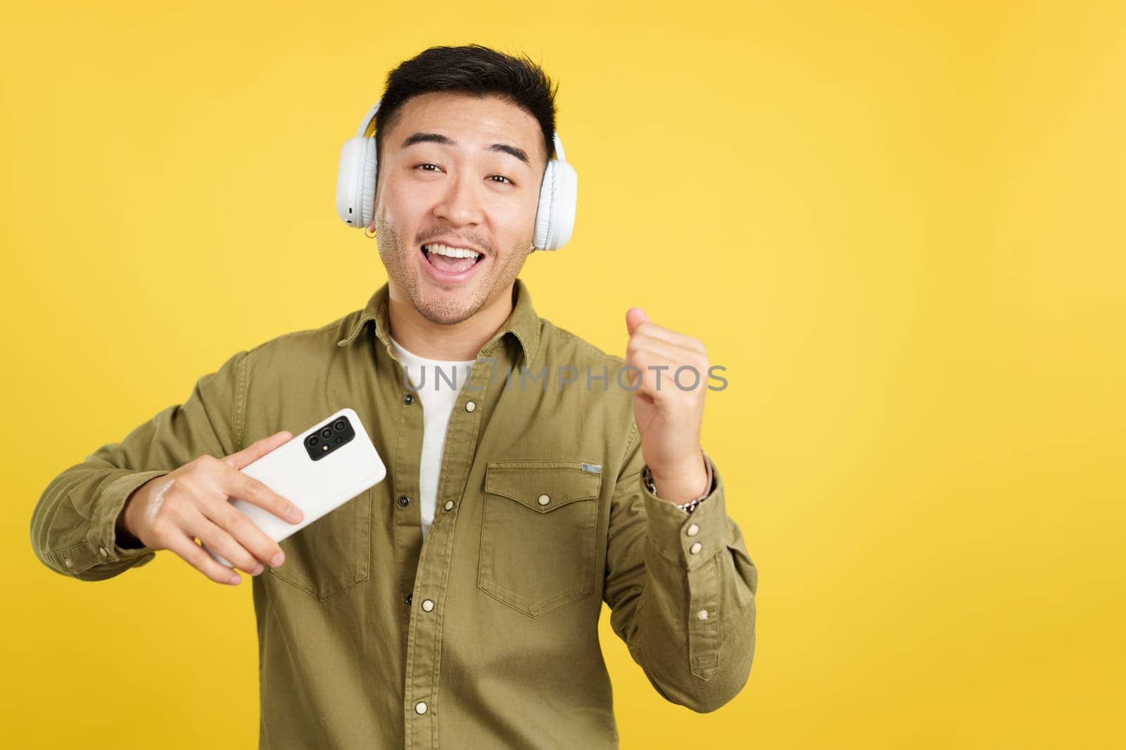 Happy chinese man dancing and listening to music with the mobile phone and headphones in studio with yellow background