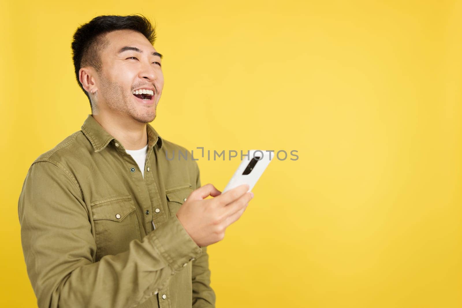 Happy chinese man laughing while using the mobile in studio with yellow background