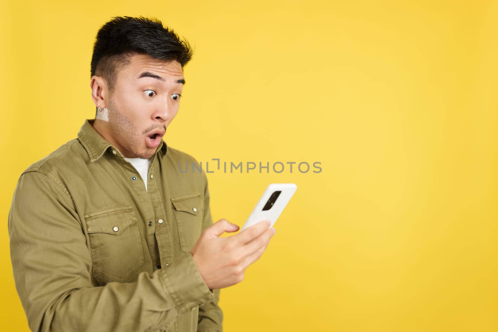 Surprised chinese man looking the screen of a mobile in studio with yellow background
