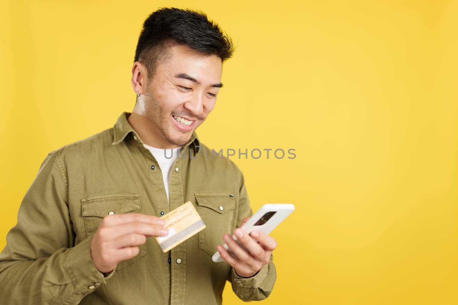Happy chinese man using the mobile and card to shopping online in studio with yellow background