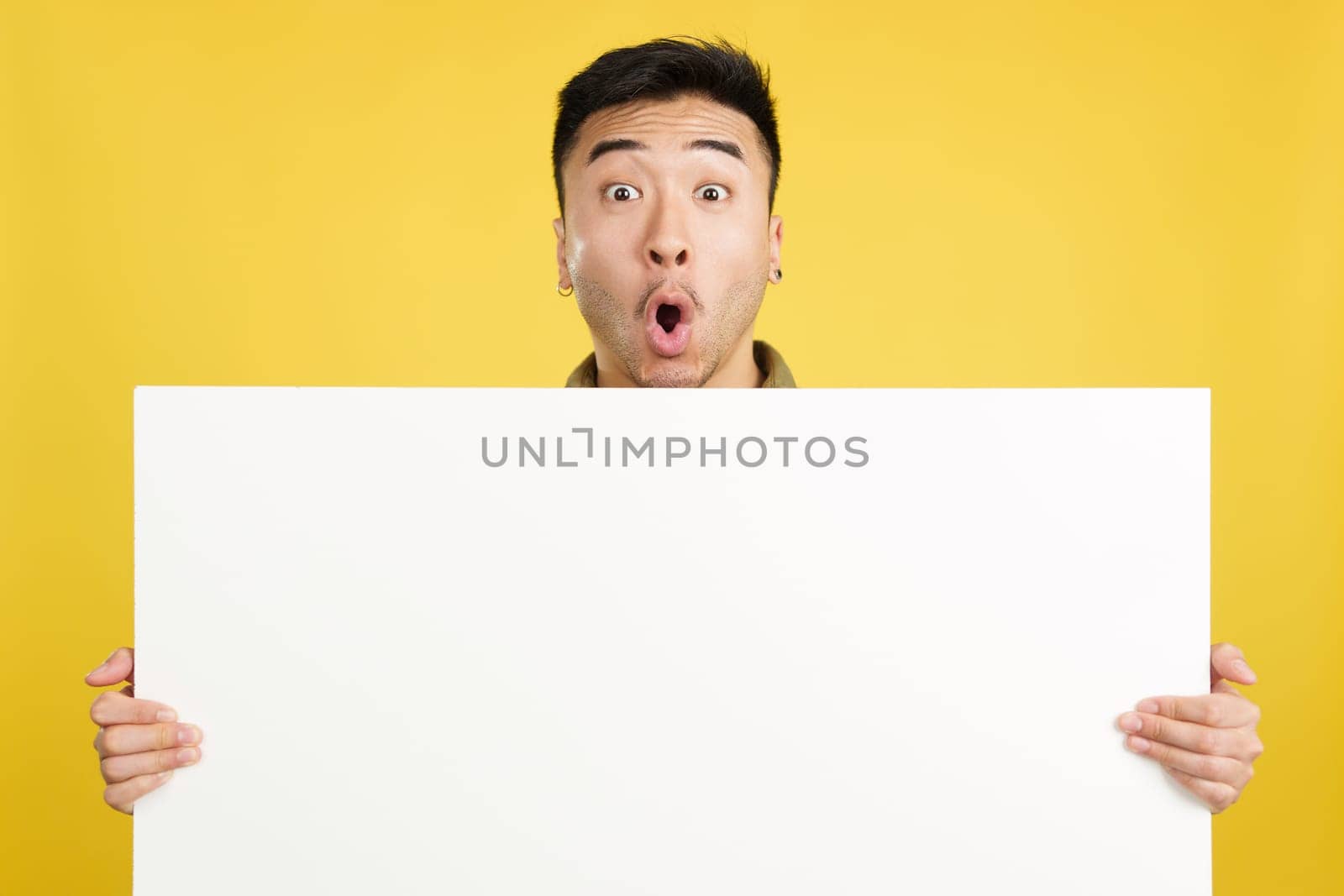 Handsome surprised chinese man hair holding a blank panel in studio with yellow background
