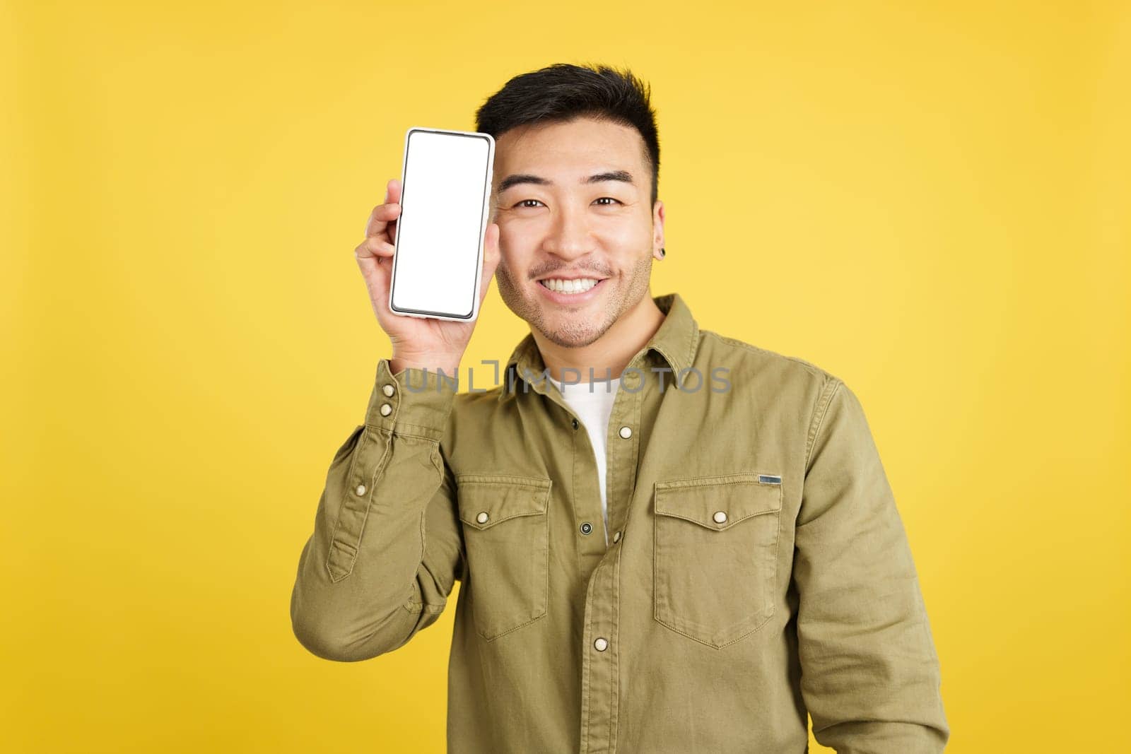 Happy chinese man looking at camera while holding a mobile with a blank screen in studio with yellow background