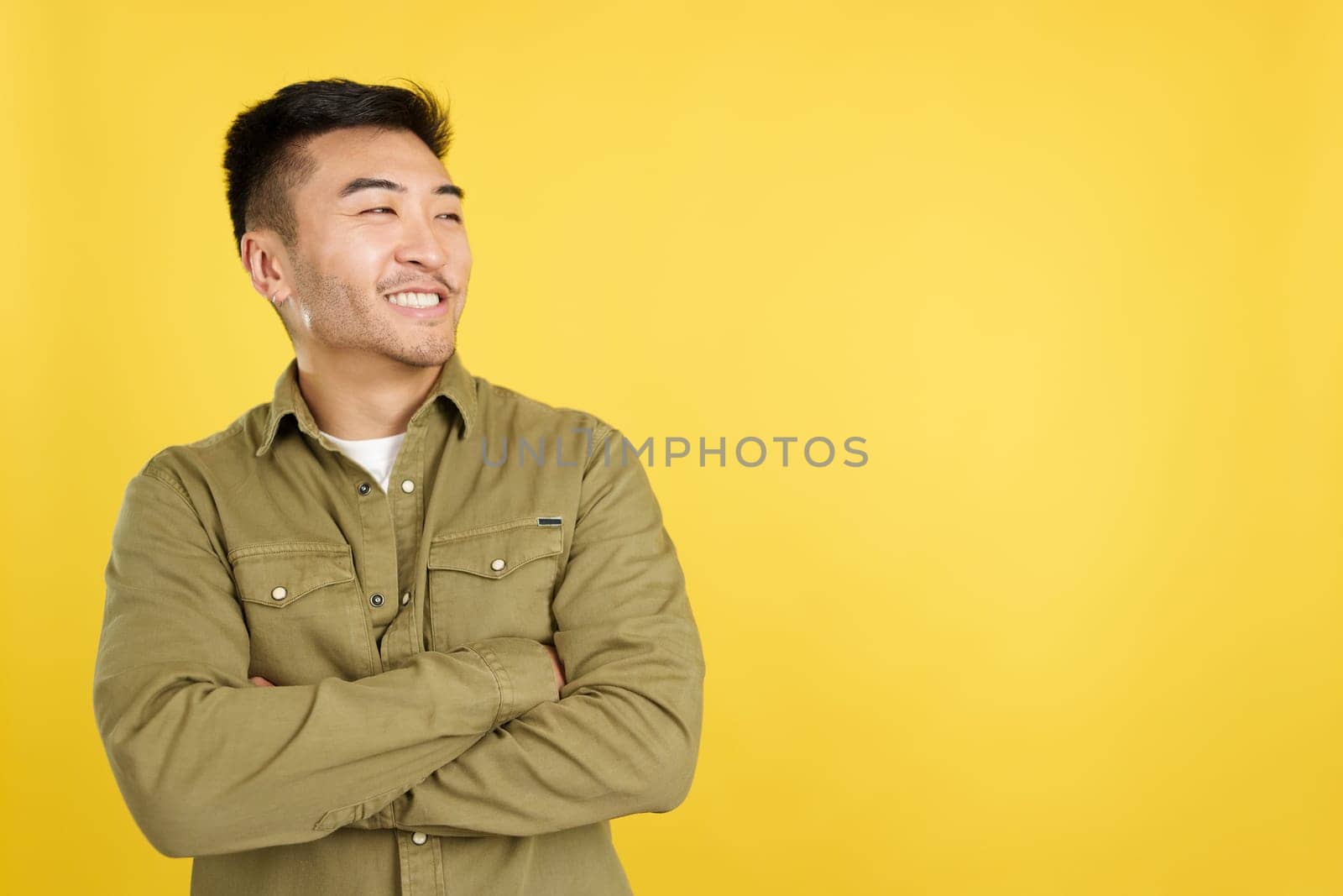 Chinese man with arms crossed smiling while looking to the side in studio with yellow background