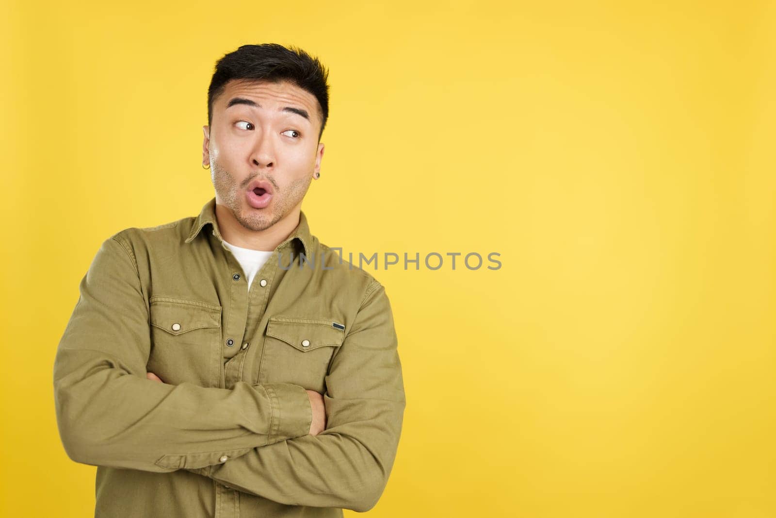 Surprised chinese man standing with open mouth and arms crossed in studio with yellow background