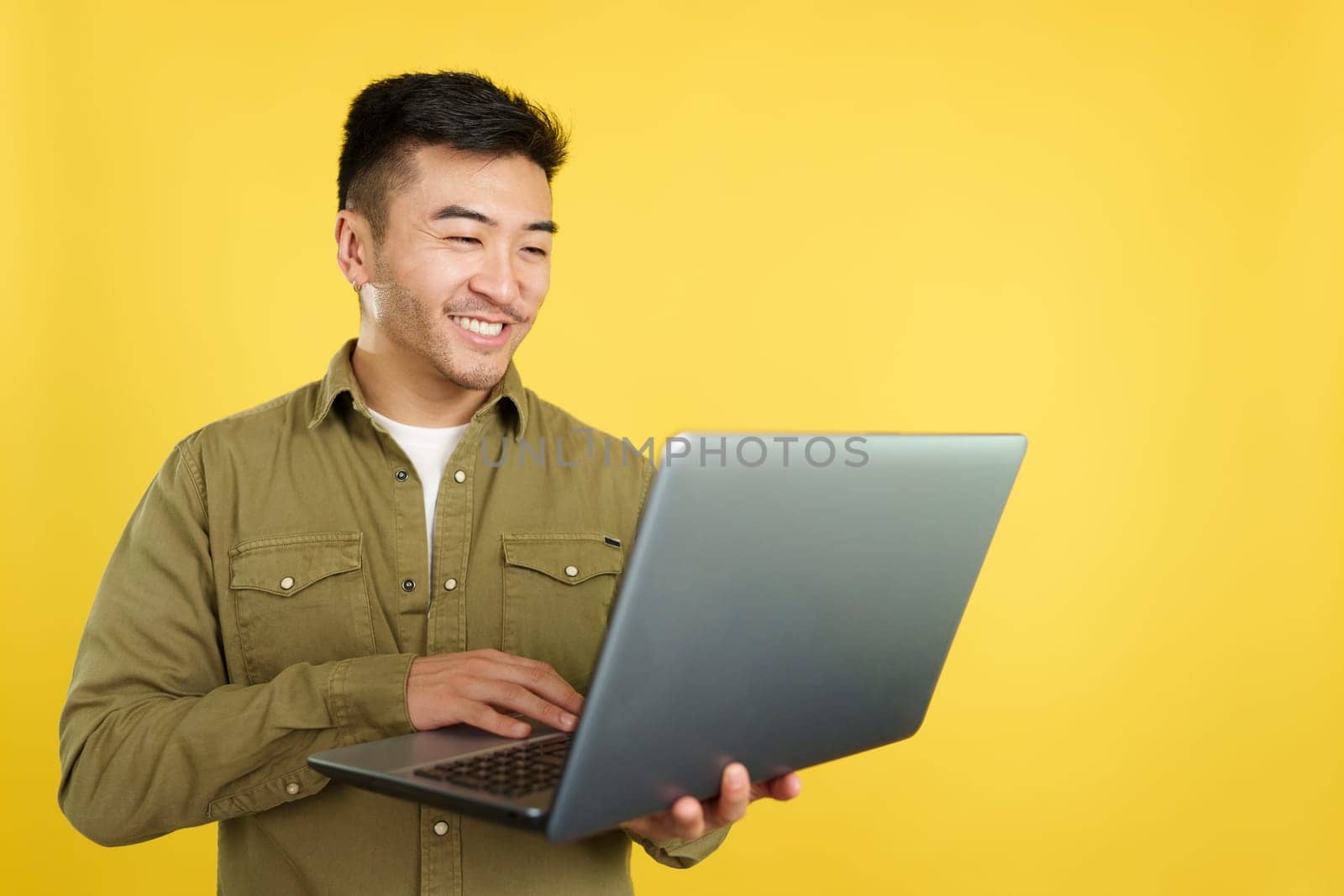 Chinese man smiling while using a laptop in studio with yellow background