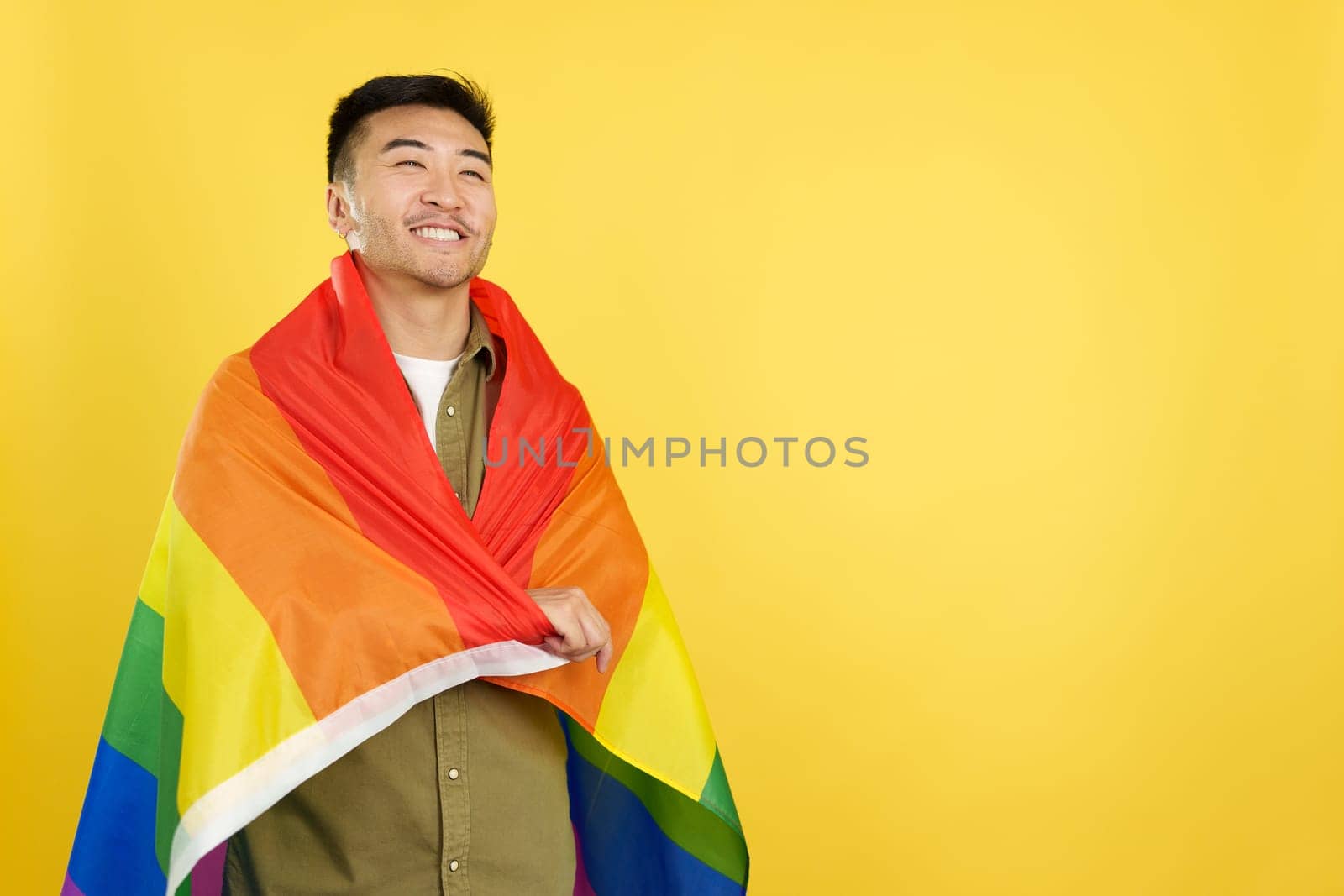 Happy chinese man wrapping with a lgbt rainbow flag in studio with yellow background