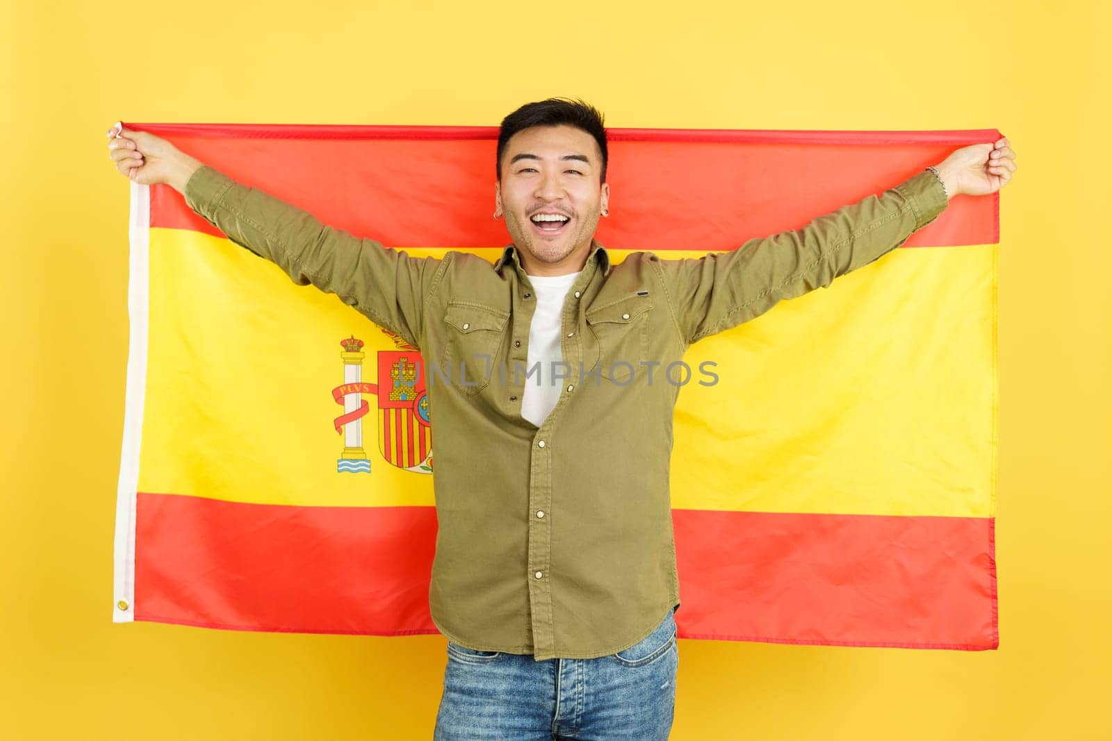 Chinese man smiling and raising a spanish national flag in studio with yellow background