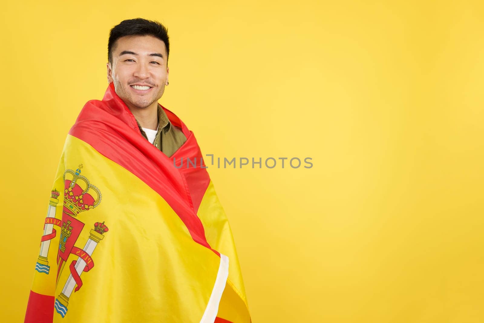 Happy chinese man looking at the camera wrapped by a spanish national flag in studio with yellow background