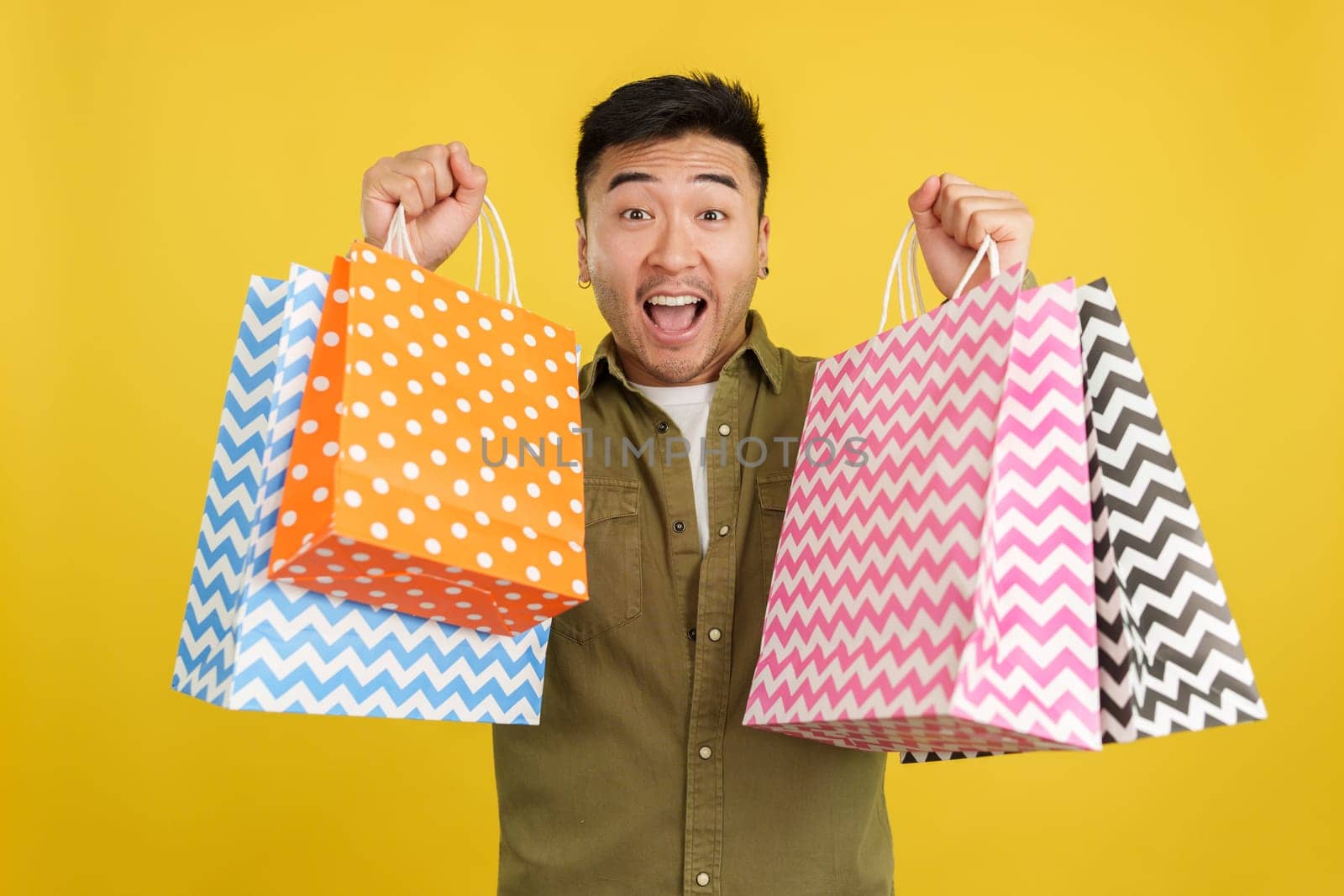 Chinese man raising many shopping bags while smiling at the camera in studio with yellow background
