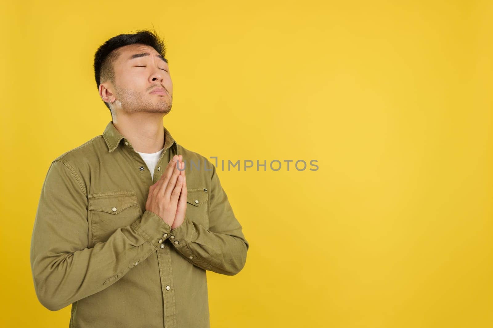 Chinese man praying with hands folded and eyes closed in studio with yellow background