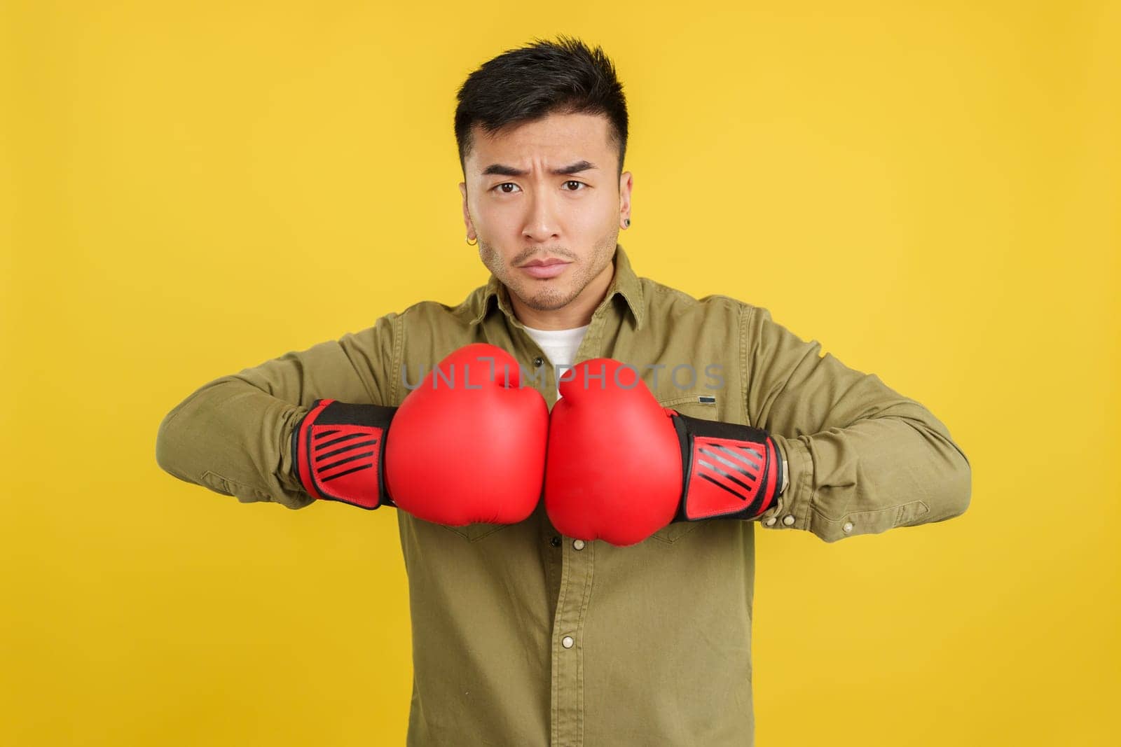 Chinese man with clashing boxing gloves while looking serious on camera in studio with yellow background