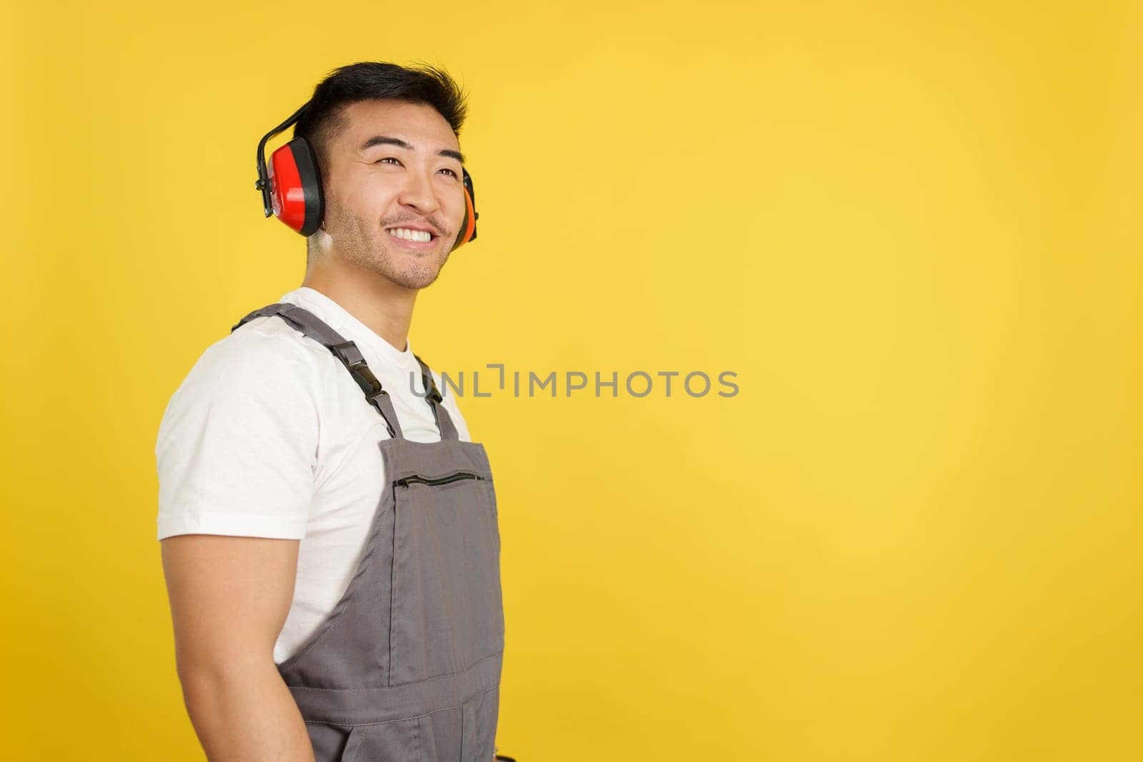 Chinese carpenter smiling distracted while looking up and wearing protective gear and headphones in studio with yellow background