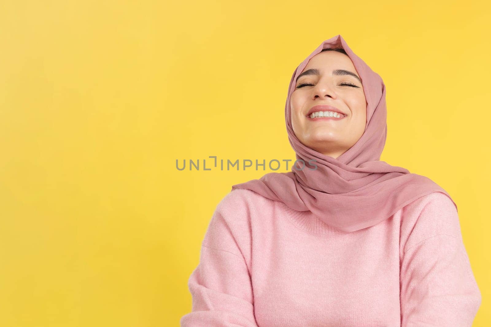 Happy muslim woman laughing with eyes closed in studio with yellow background