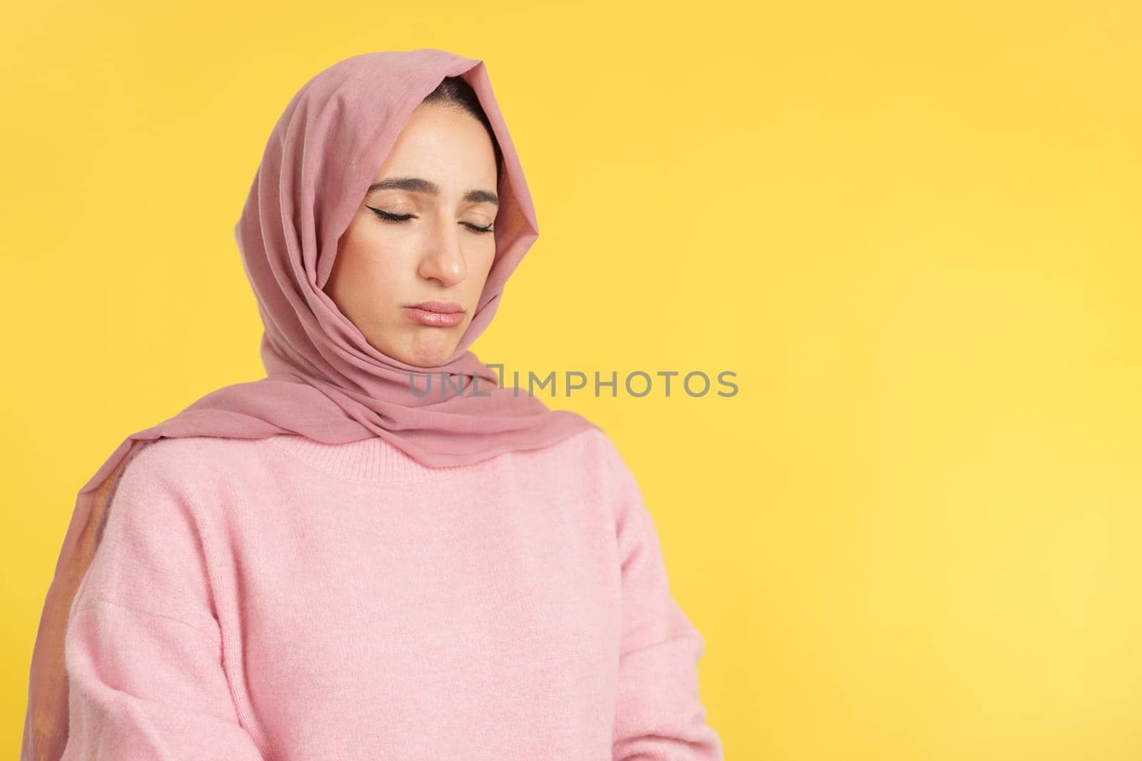 Sad muslim woman standing with eyes closed in studio with yellow background