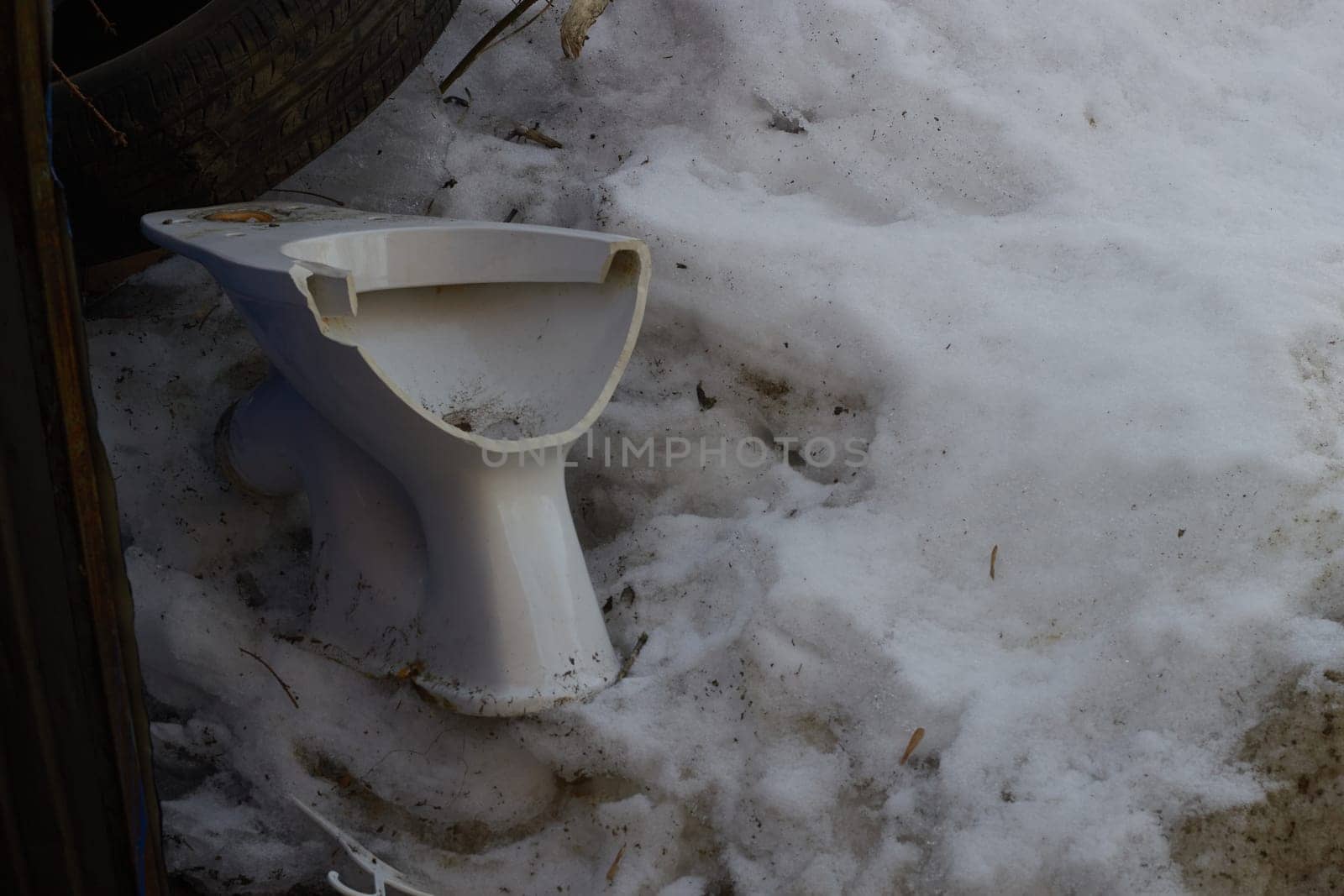 A broken white toilet bowl against a background of dirty snow. Garbage dump in the trash. Pollution. Sorting garbage.