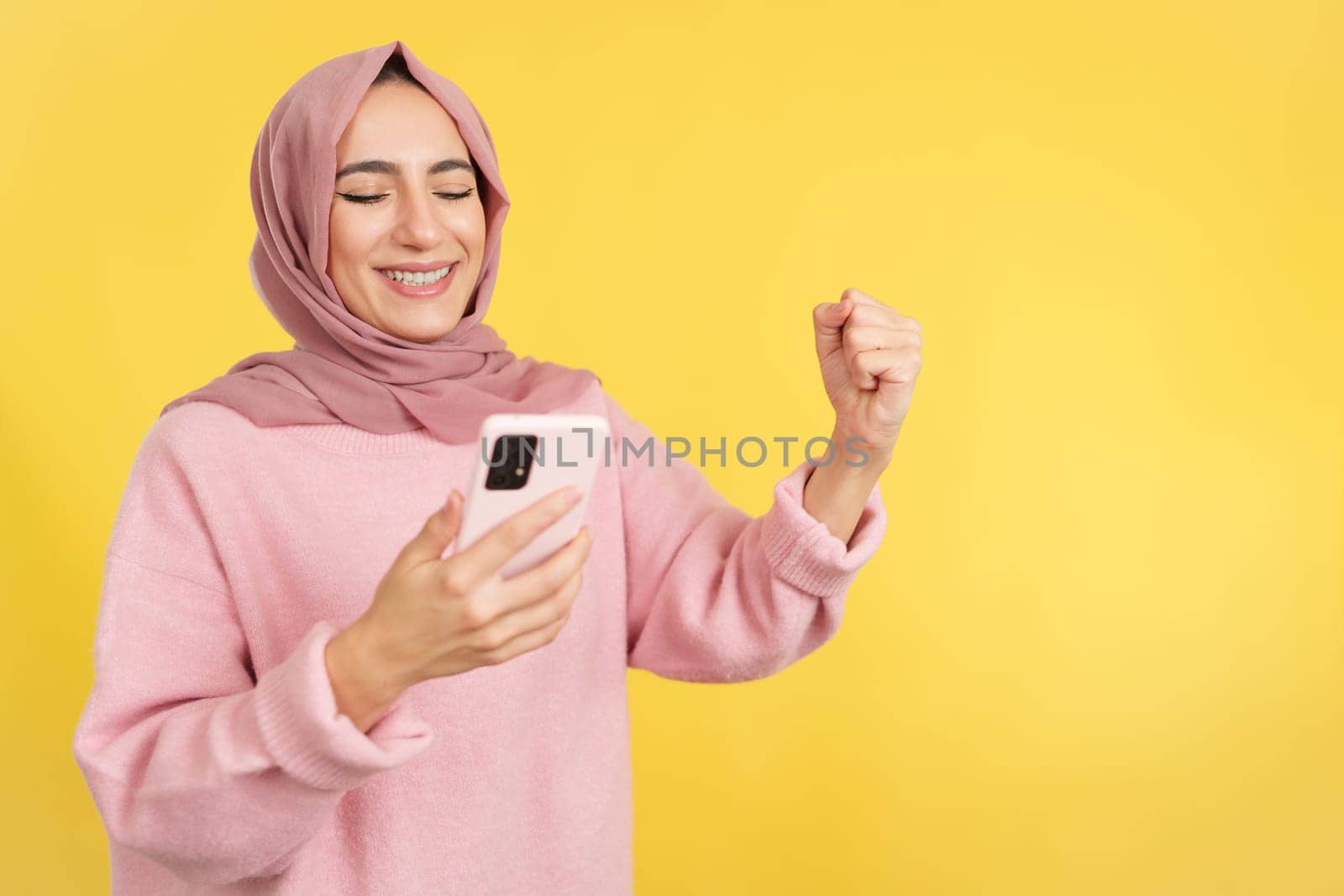 Excited muslim woman celebrating raising the fist while using a mobile in studio with yellow background
