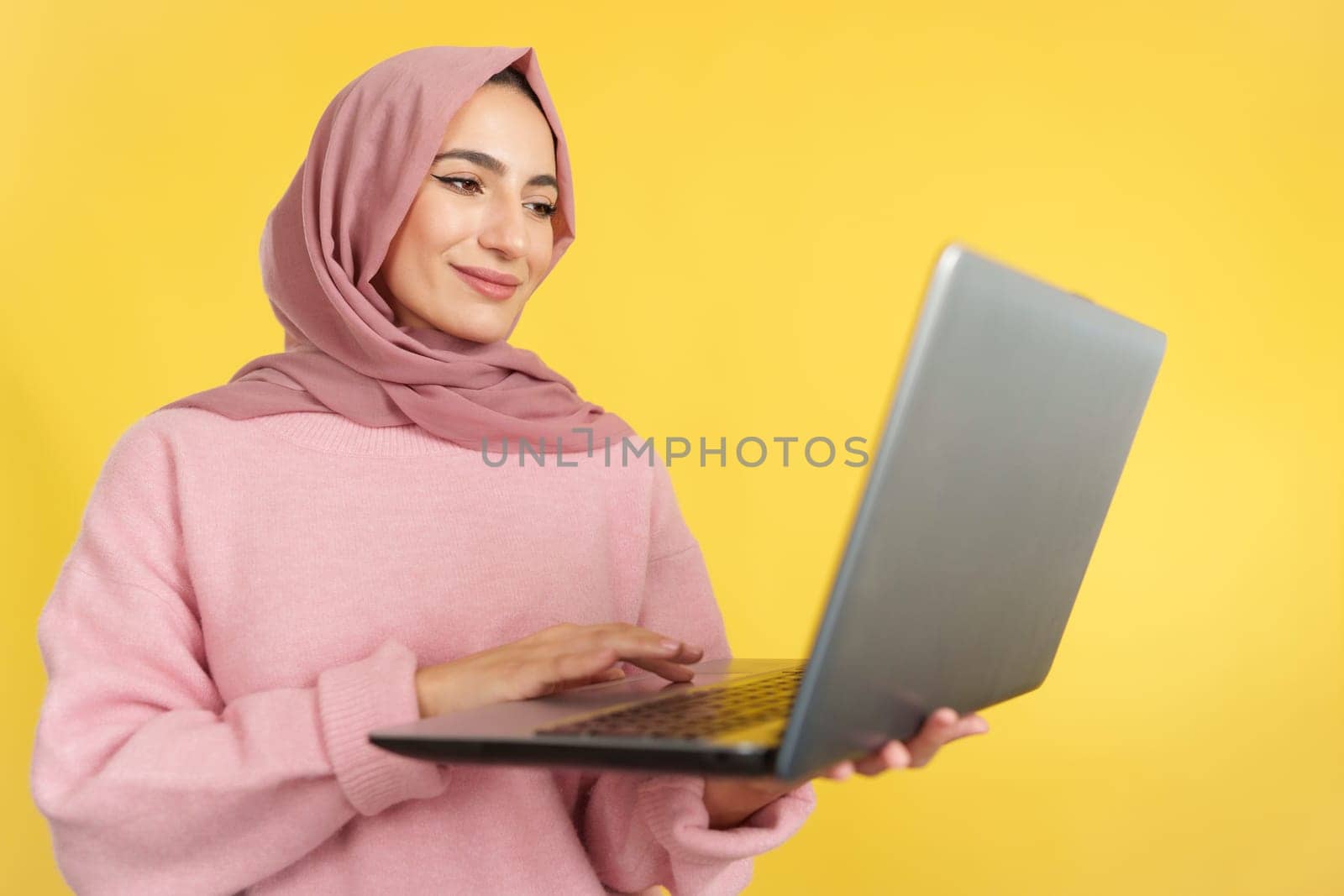 Happy muslim woman standing using a laptop in studio with yellow background