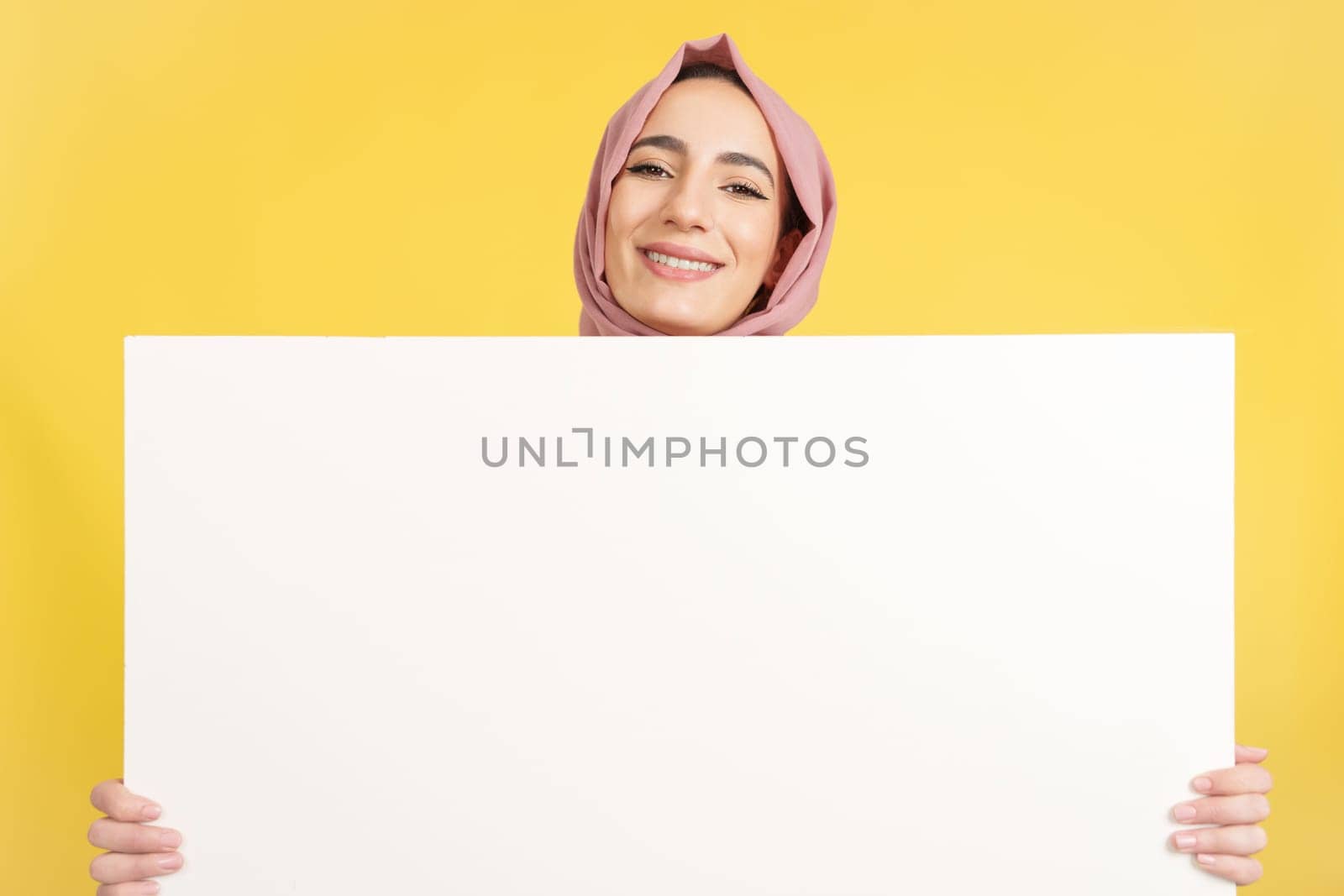 Happy muslim woman with scarf holding a blank panel in studio with yellow background