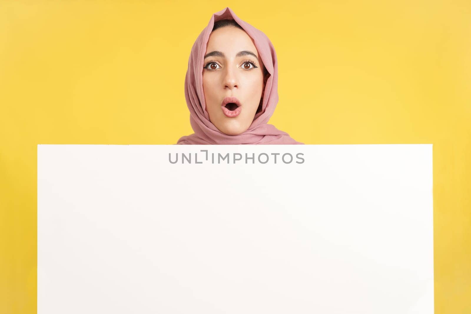Surprised muslim woman with scarf holding a blank panel in studio with yellow background