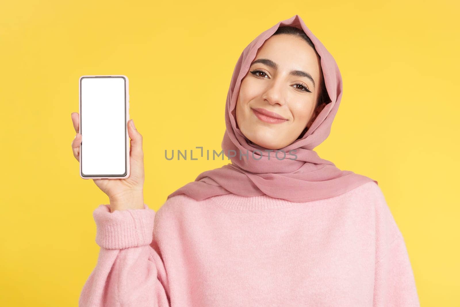 Happy muslim woman holding a mobile with a blank screen in studio with yellow background