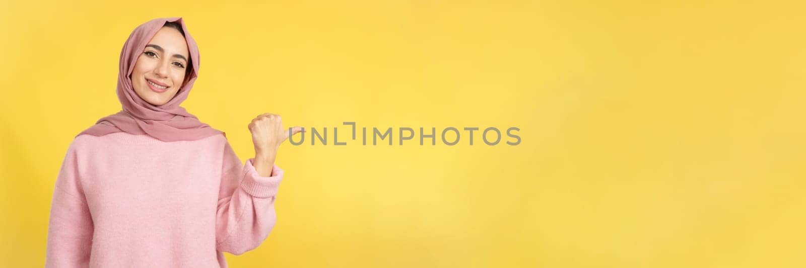 Muslim woman smiling at camera and pointing to the side in studio with yellow background