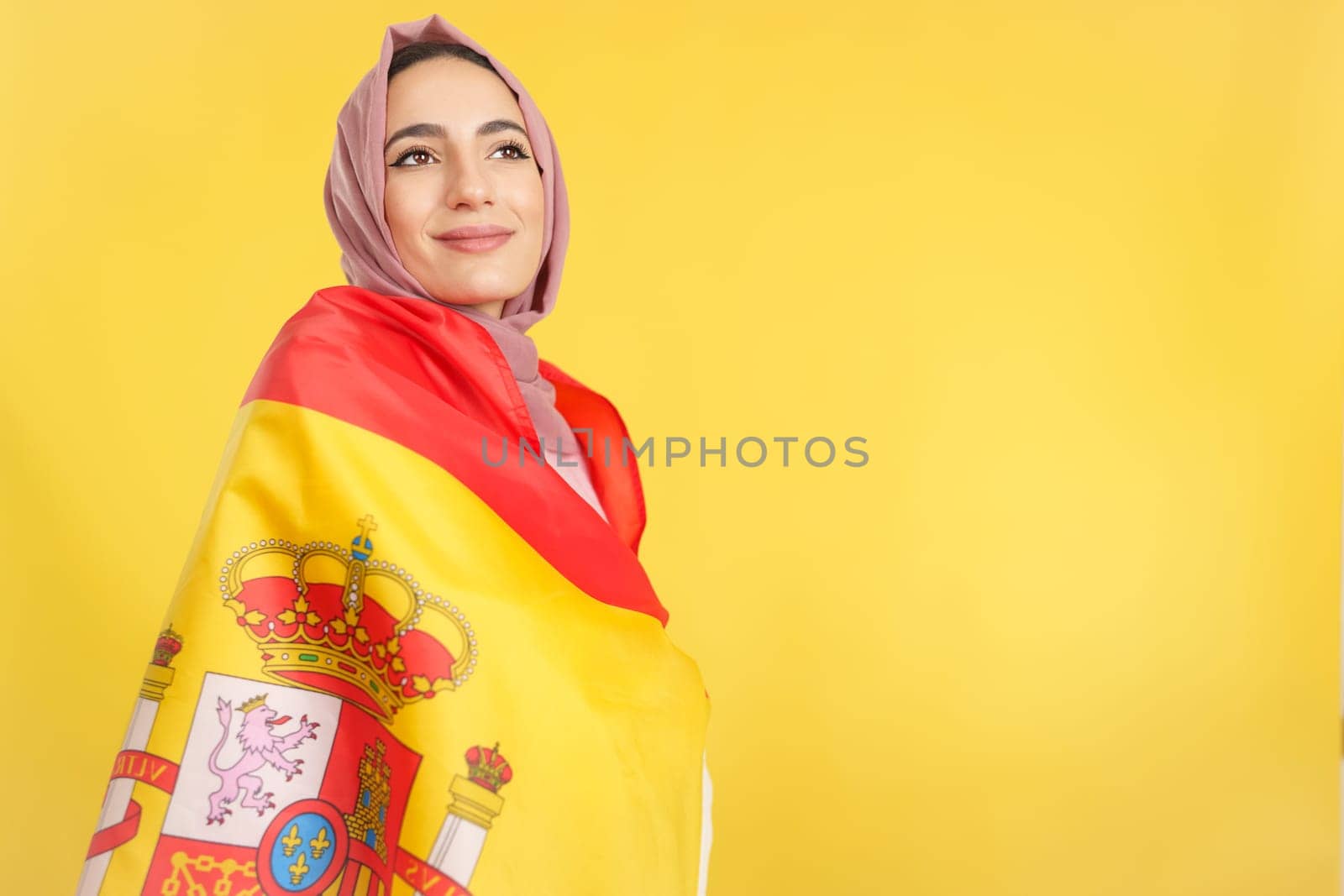 Dreamy muslim woman wrapped by a spanish national flag in studio with yellow background
