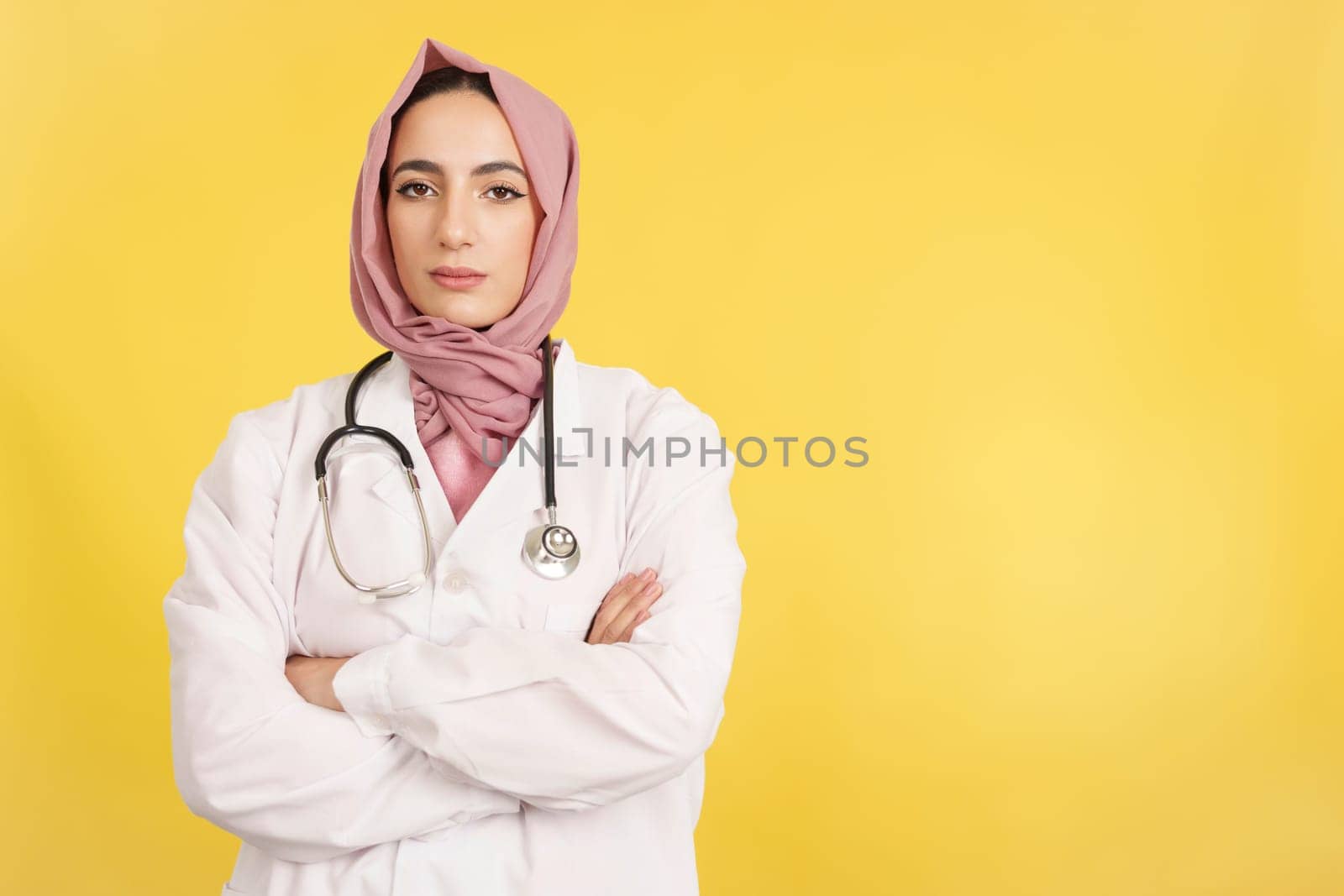 Serious female muslim doctor with arms crossed looking at camera in studio with yellow background