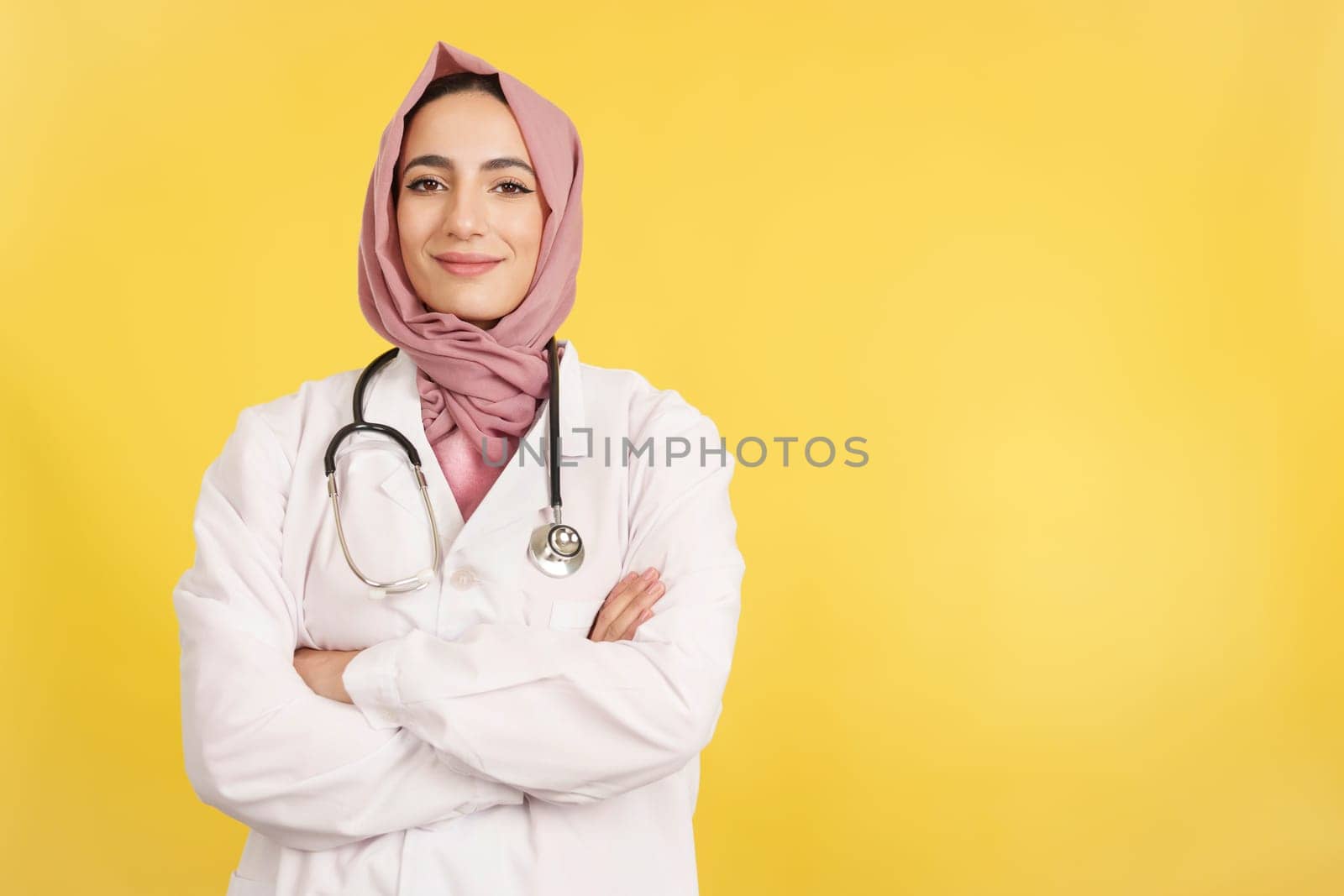 Happy female muslim doctor with arms crossed looking at camera in studio with yellow background