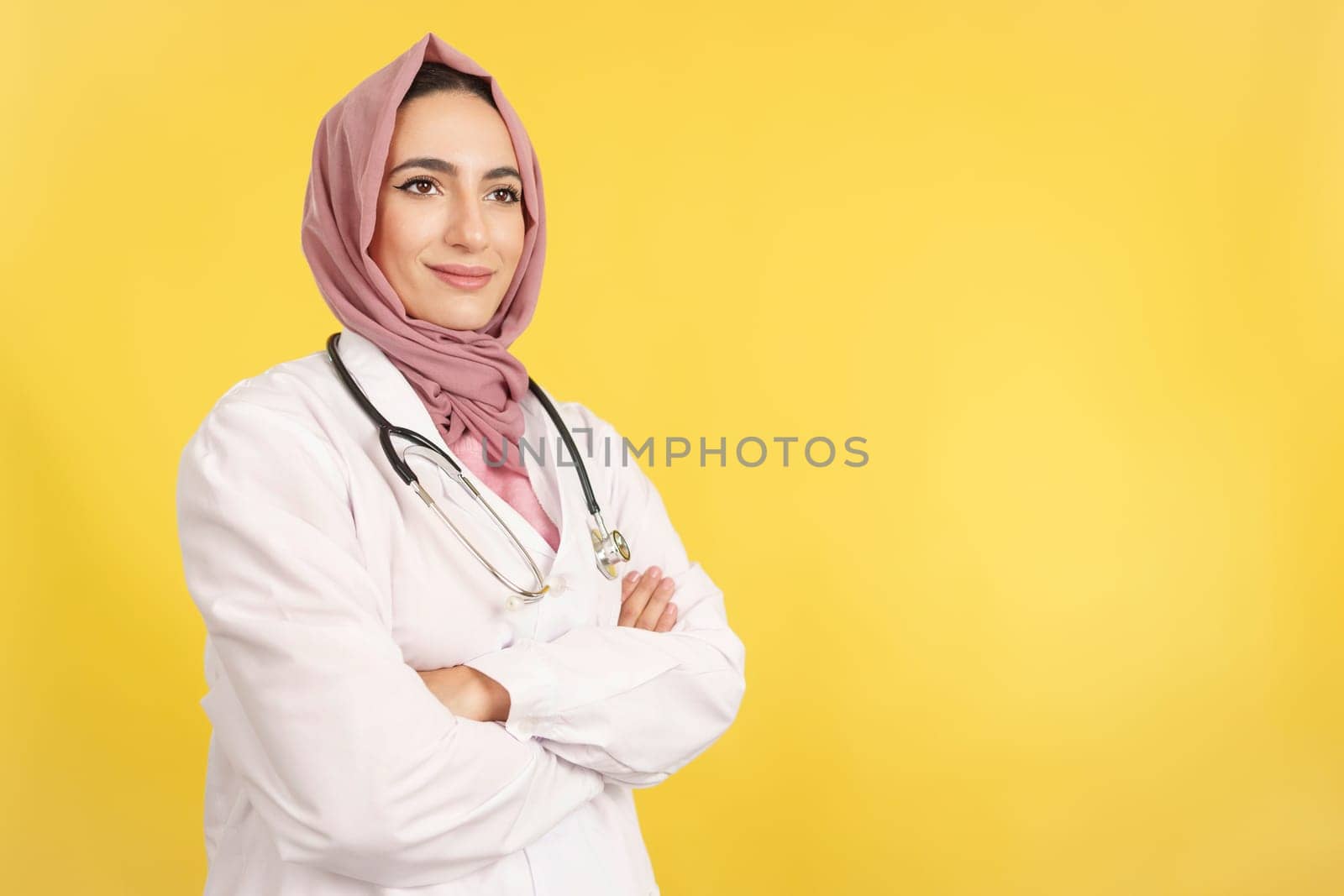 Happy female muslim doctor with arms crossed looking away in studio with yellow background