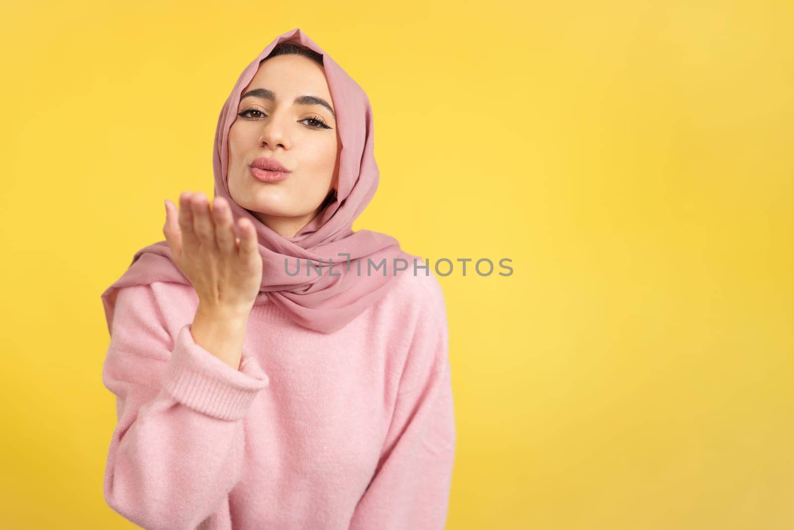 Muslim woman blowing a kiss while looking at the camera in studio with yellow background