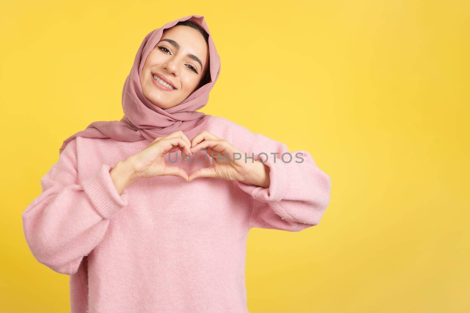 Muslim woman representing a heart shape of fingers in studio with yellow background