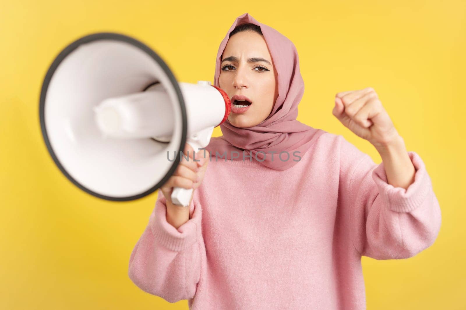 Angry muslim woman screaming using a loudspeaker in studio with yellow background