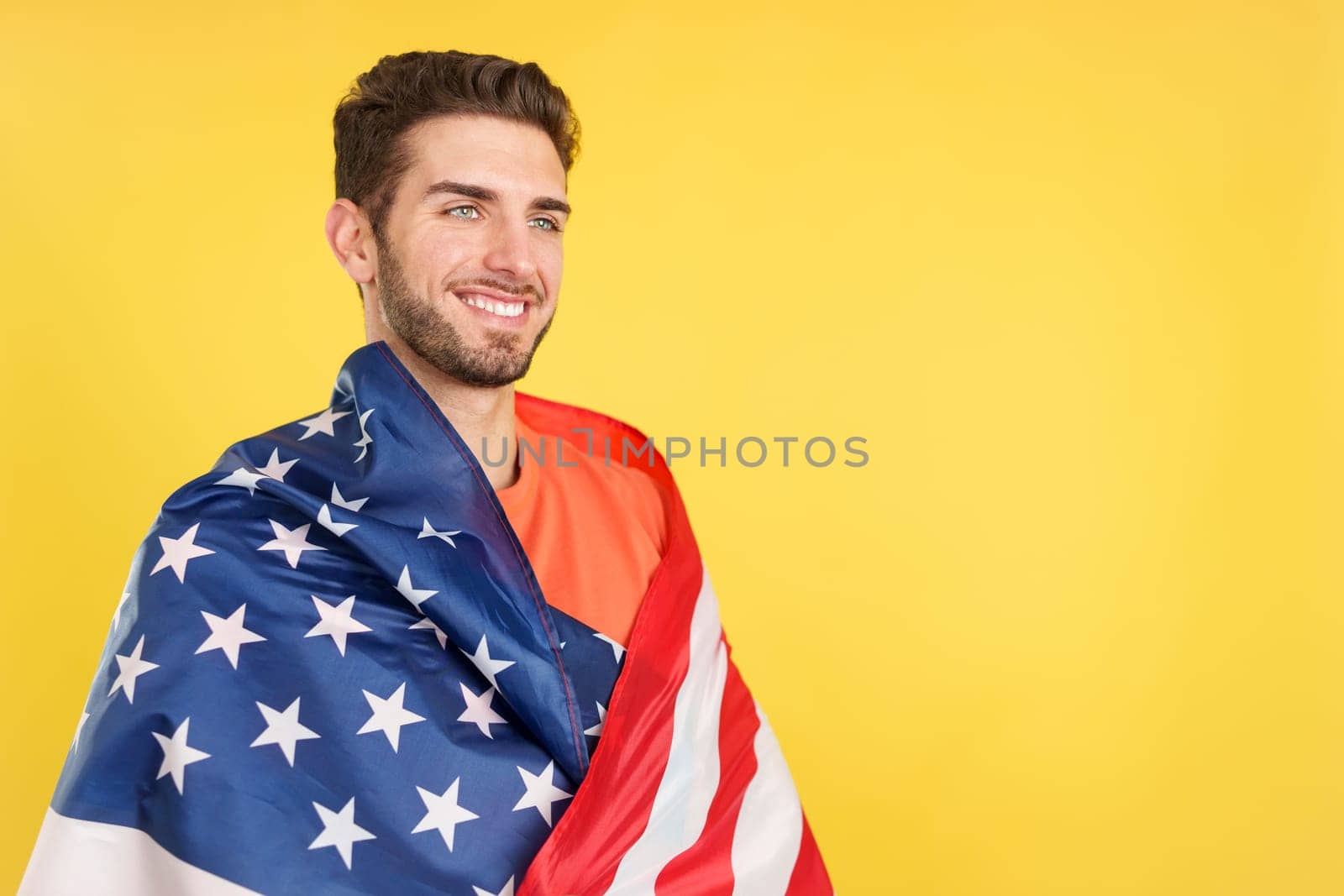 Proud caucasian man wrapping with a north america national flag in studio with yellow background