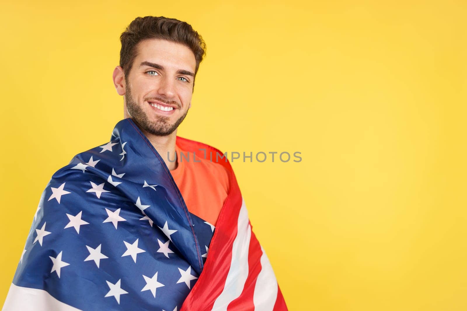 Happy caucasian man wrapping with a north america national flag in studio with yellow background