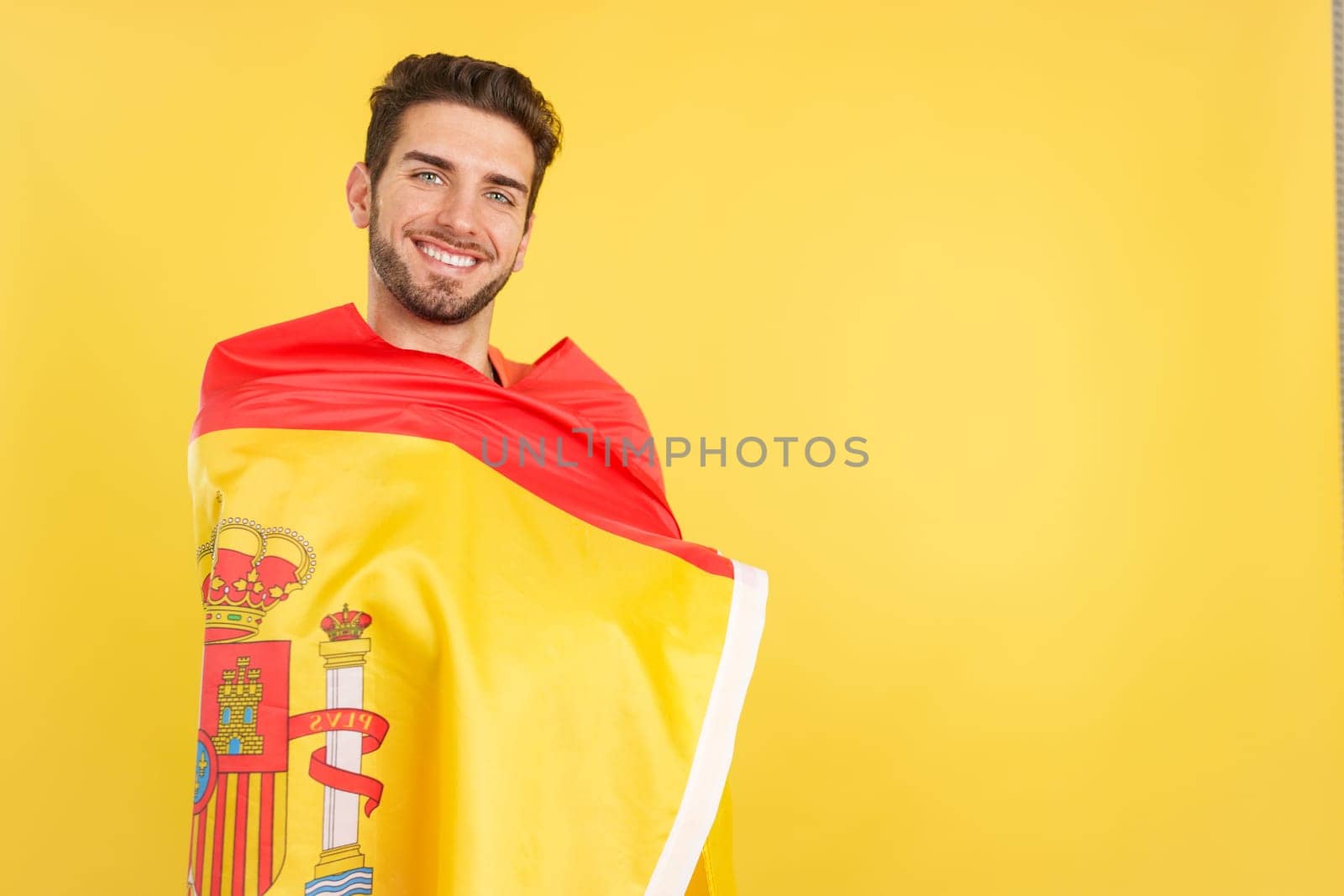 Happy caucasian man wrapping with a spanish national flag in studio with yellow background