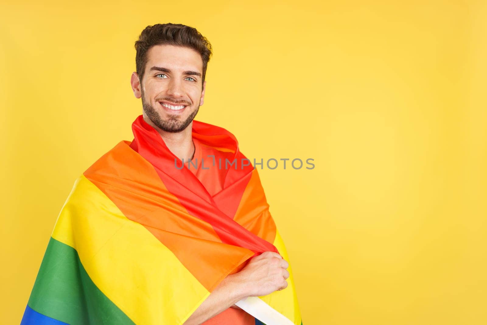 Happy caucasian man wrapped with a lgbt flag in studio with yellow background