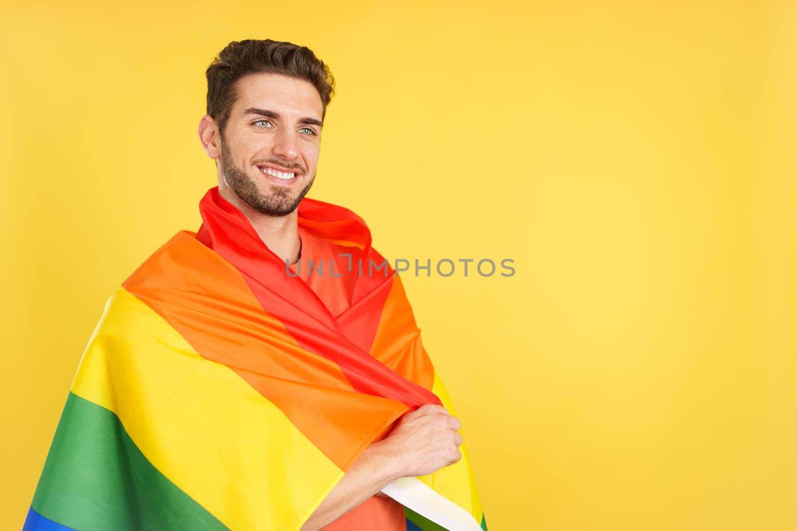 Distracted caucasian man wrapped with a lgbt flag in studio with yellow background