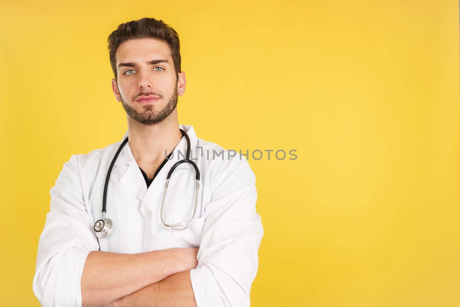 Serious caucasian doctor looking at the camera with arms crossed in studio with yellow background