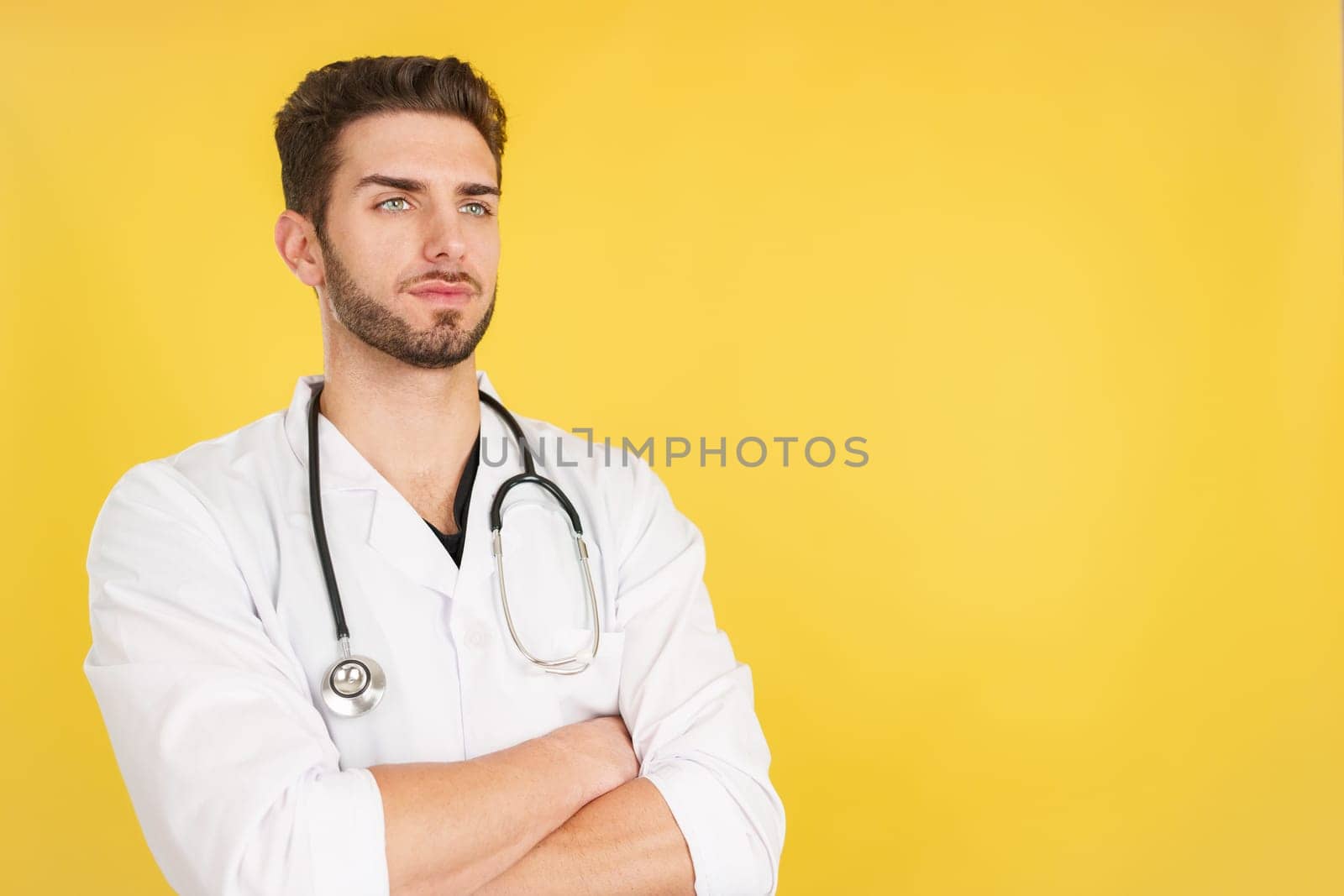 Serious caucasian doctor with arms crossed looking away in studio with yellow background