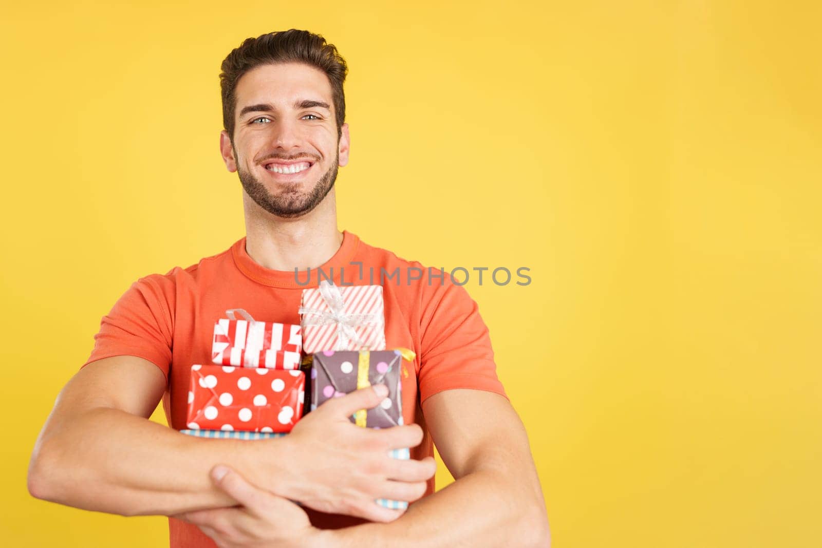 Happy caucasian man holing many colorful gifts in studio with yellow background