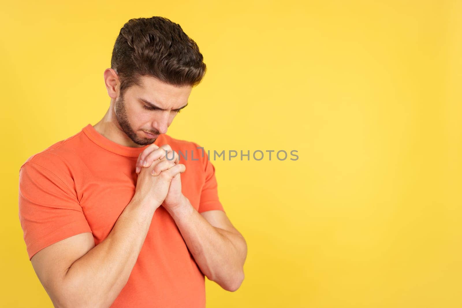 Caucasian man praying with folded hands looking down in studio with yellow background