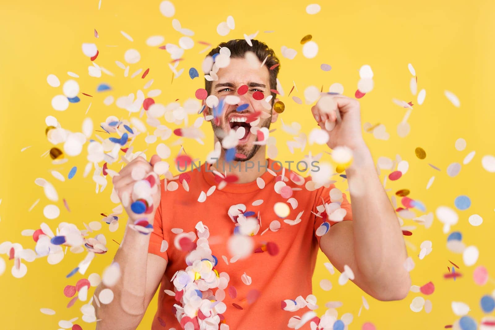 Happy caucasian man surrounded by confetti flying in the air in studio with yellow background