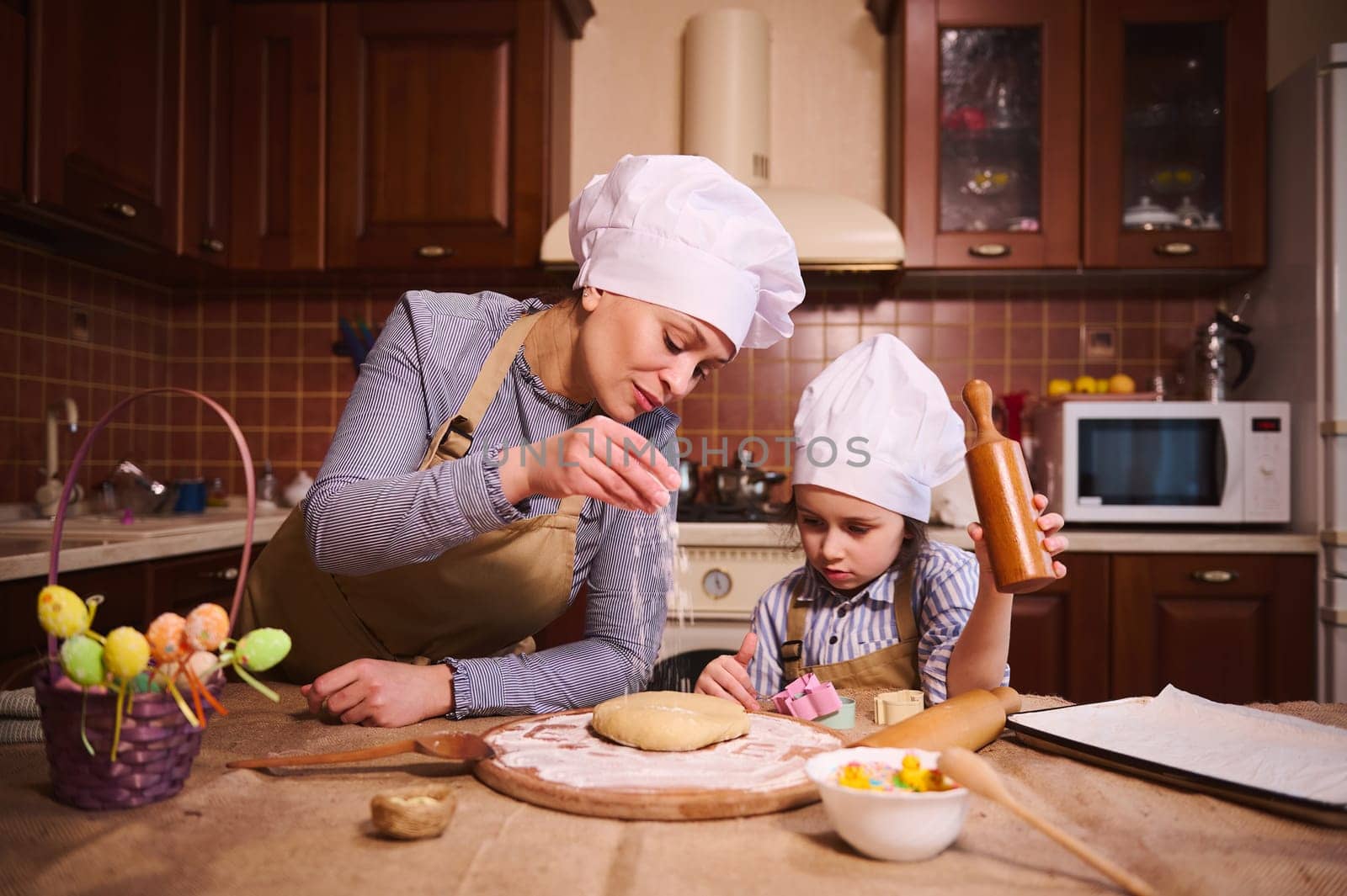 Multi-ethnic pregnant pretty woman, loving mom in chef's hat and beige apron, standing with her lovely daughter at kitchen island, flouring dough, preparing cakes and panettone for Easter holiday