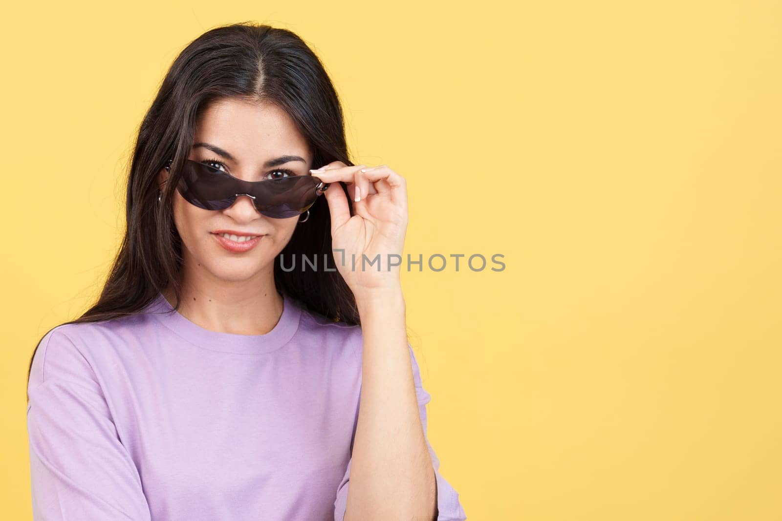 Hispanic woman lowering her sunglasses to see and look at the camera in studio with yellow background