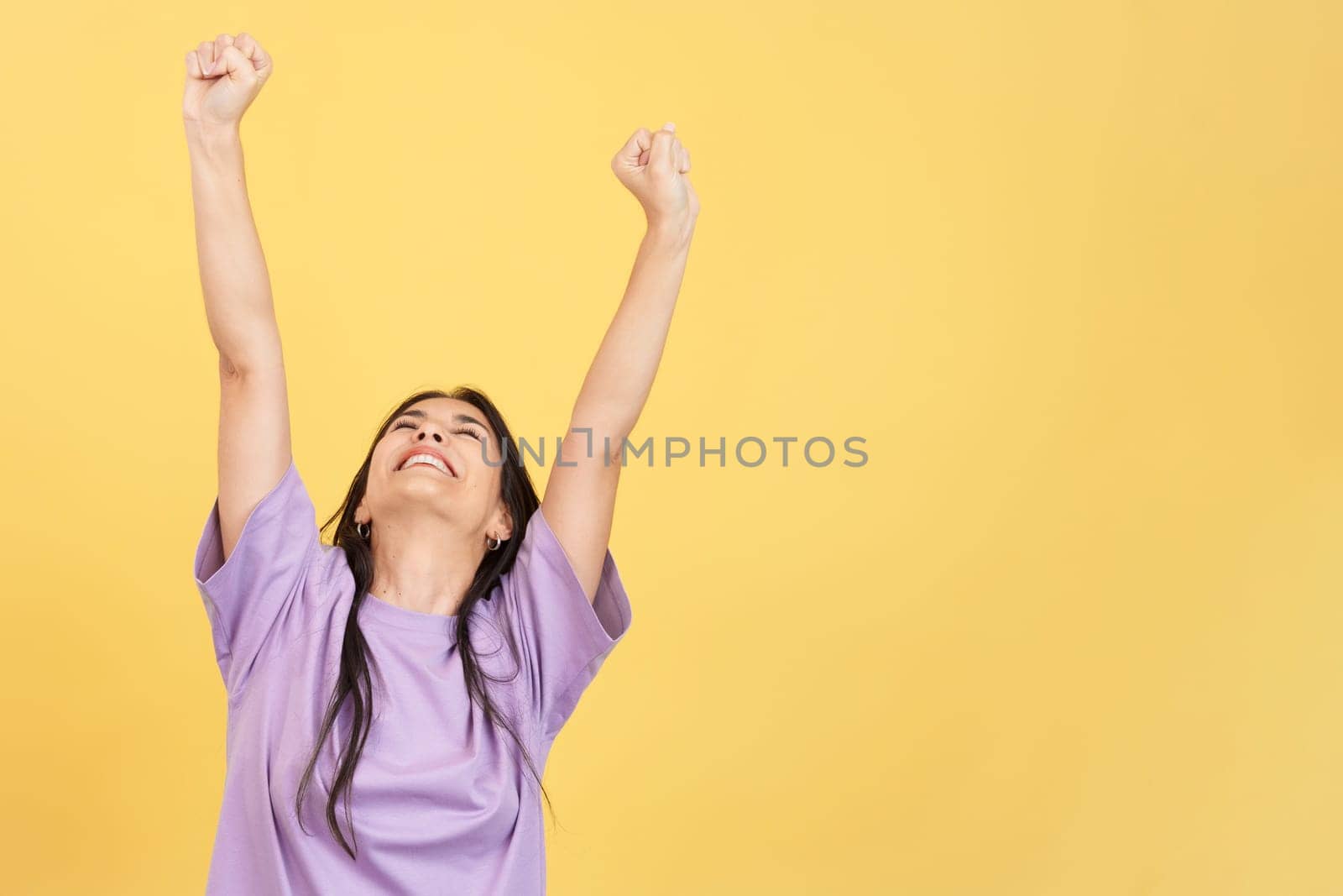 Happy hispanic woman celebrating while raising arms in studio with yellow background
