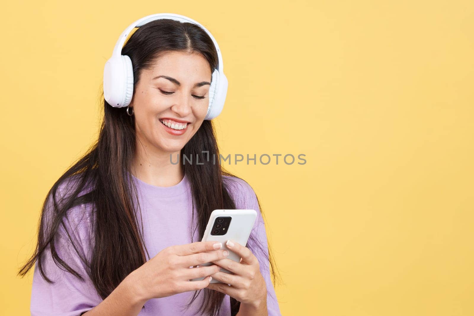Caucasian woman listening to music with headphones and a mobile in studio with yellow background
