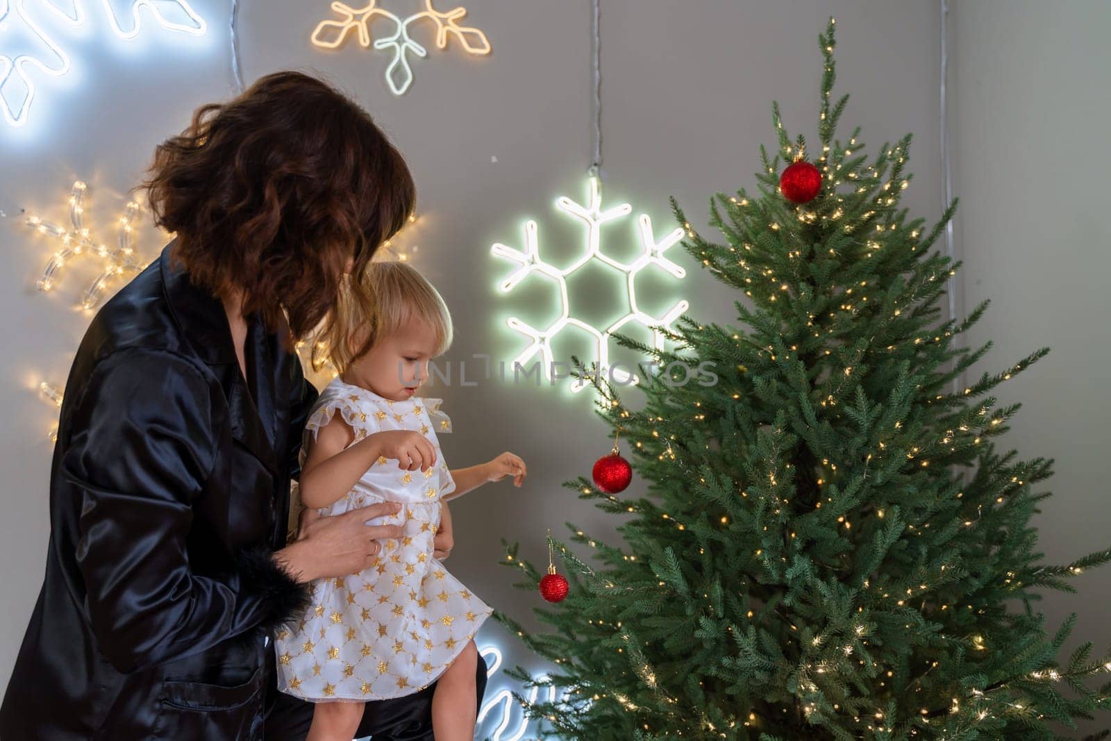 A mother with a 2-year-old daughter decorates the Christmas tree. Mom in a black suit, a girl in a white dress, her daughter hangs a red ball on the Christmas tree. Merry Christmas and New Year concept by Matiunina
