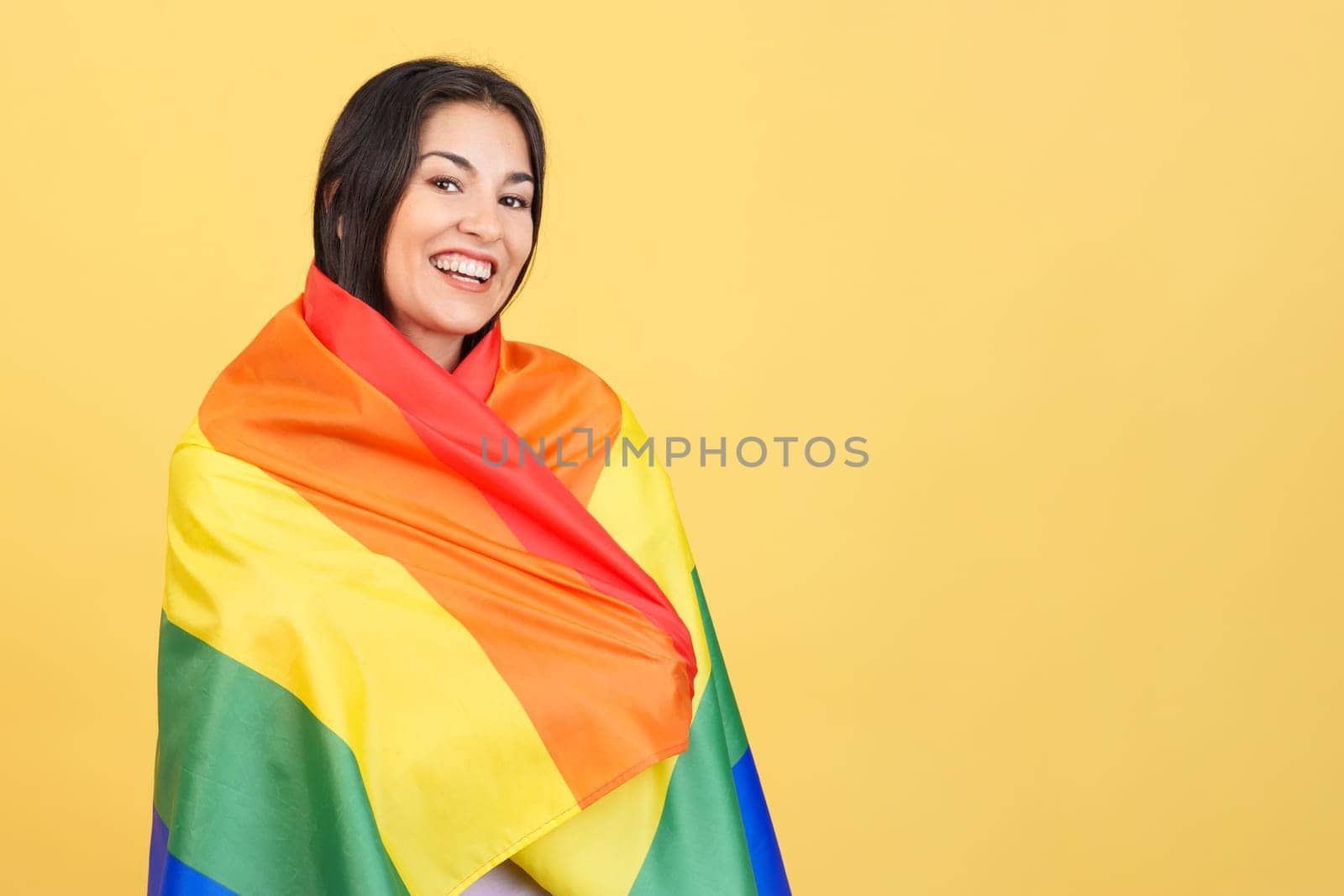 Happy caucasian woman wrapped with a lgbt flag in studio with yellow background