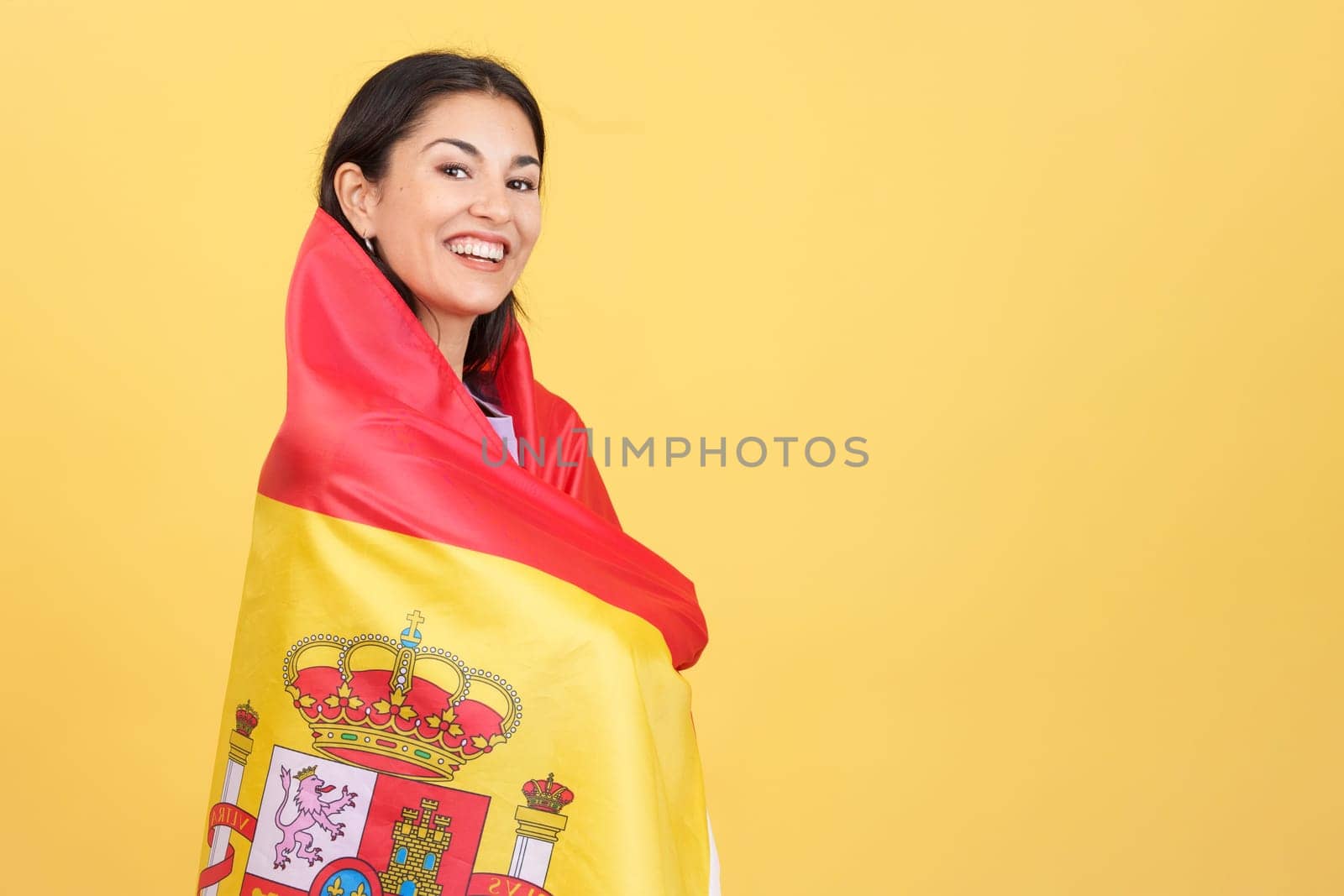 Happy caucasian woman wrapping with a spanish national flag in studio with yellow background